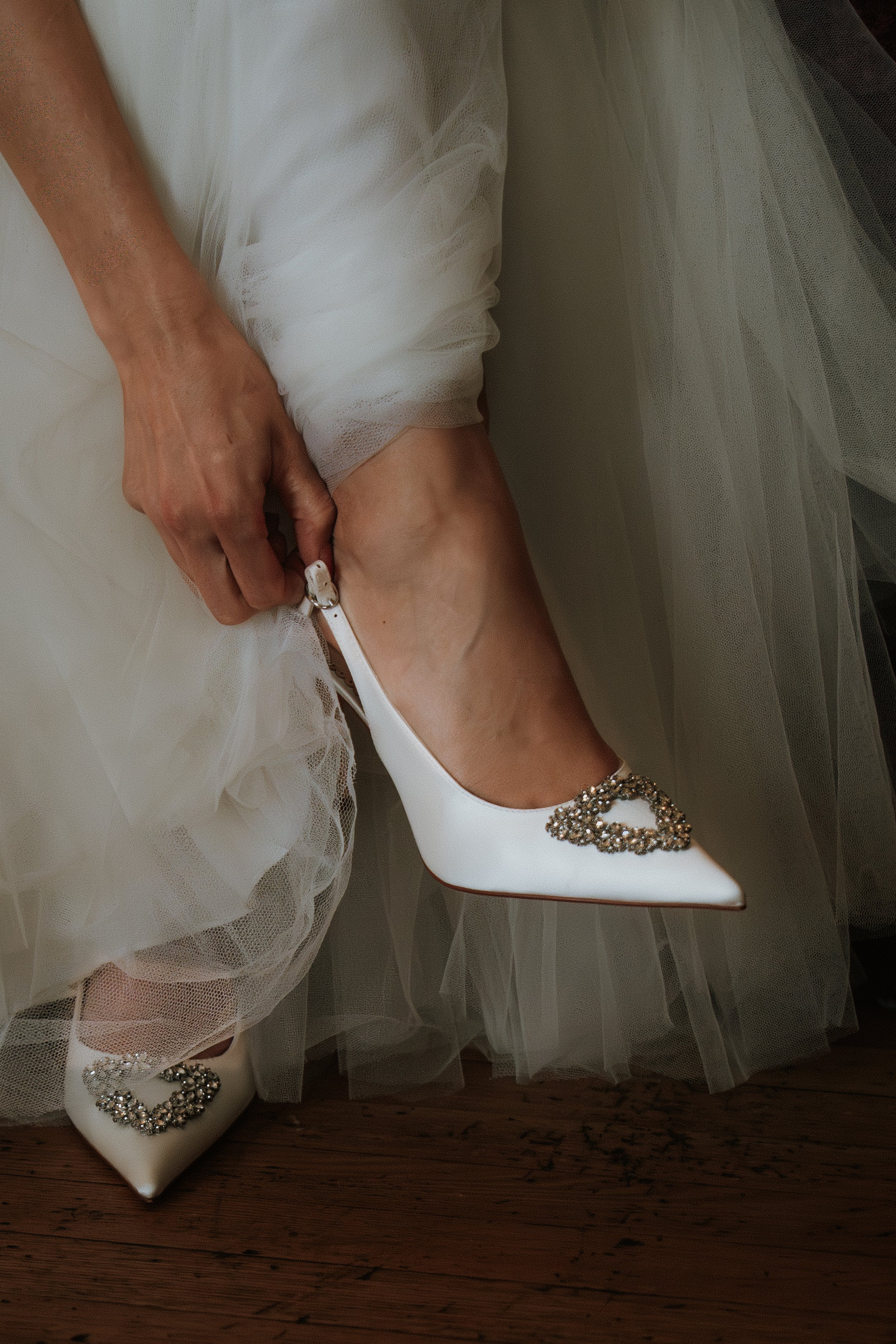 Close-up bridal detail shot of a woman wearing iconic white Manolo Blahnik wedding shoes with rhinestone embellishments, stepping out from beneath a luxury tulle bridal gown.