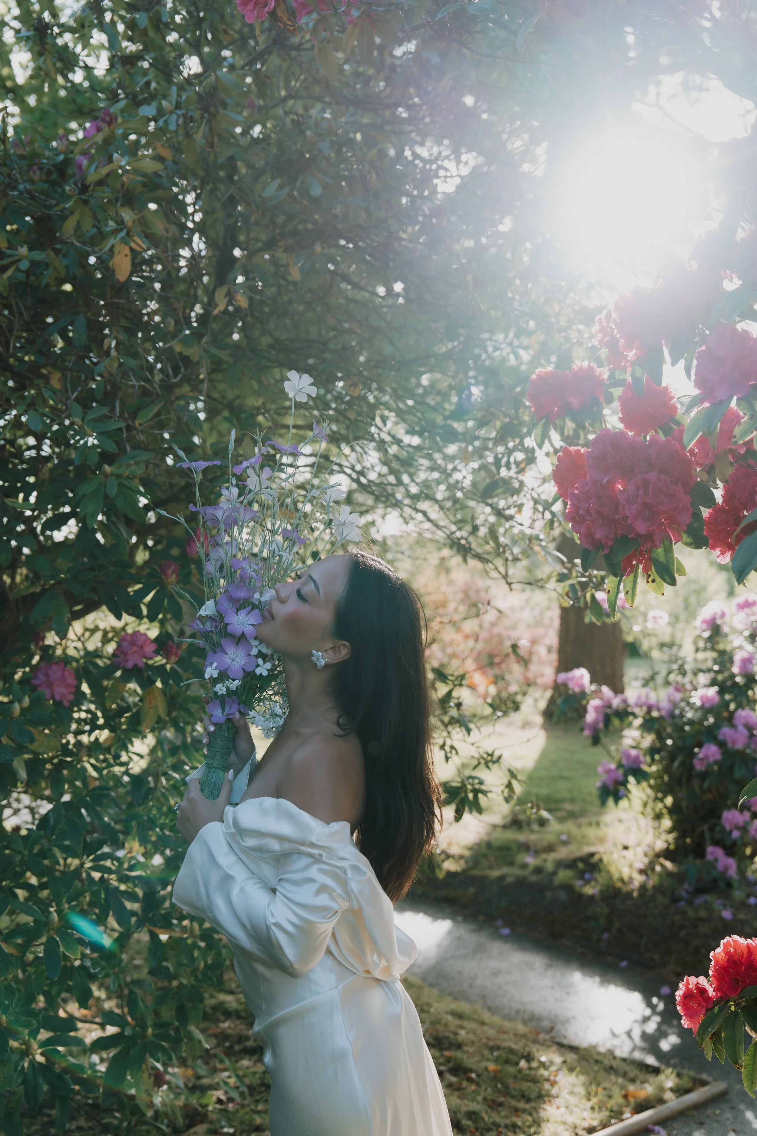 A Danielle Frankel bride wearing a romantic off-the-shoulder wedding dress and holding a purple and white bouquet, standing outdoors under sunlit tree branches filled with blossoms.