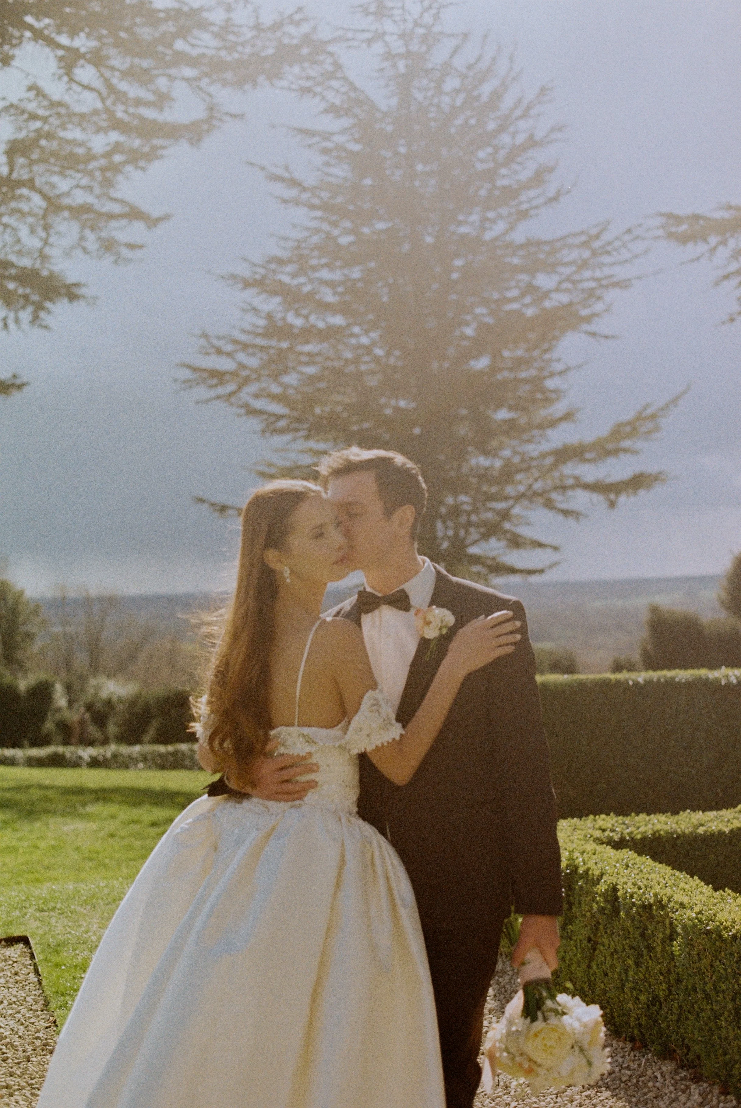 A bride and groom embrace outdoors, with a large tree and manicured bushes in the background.