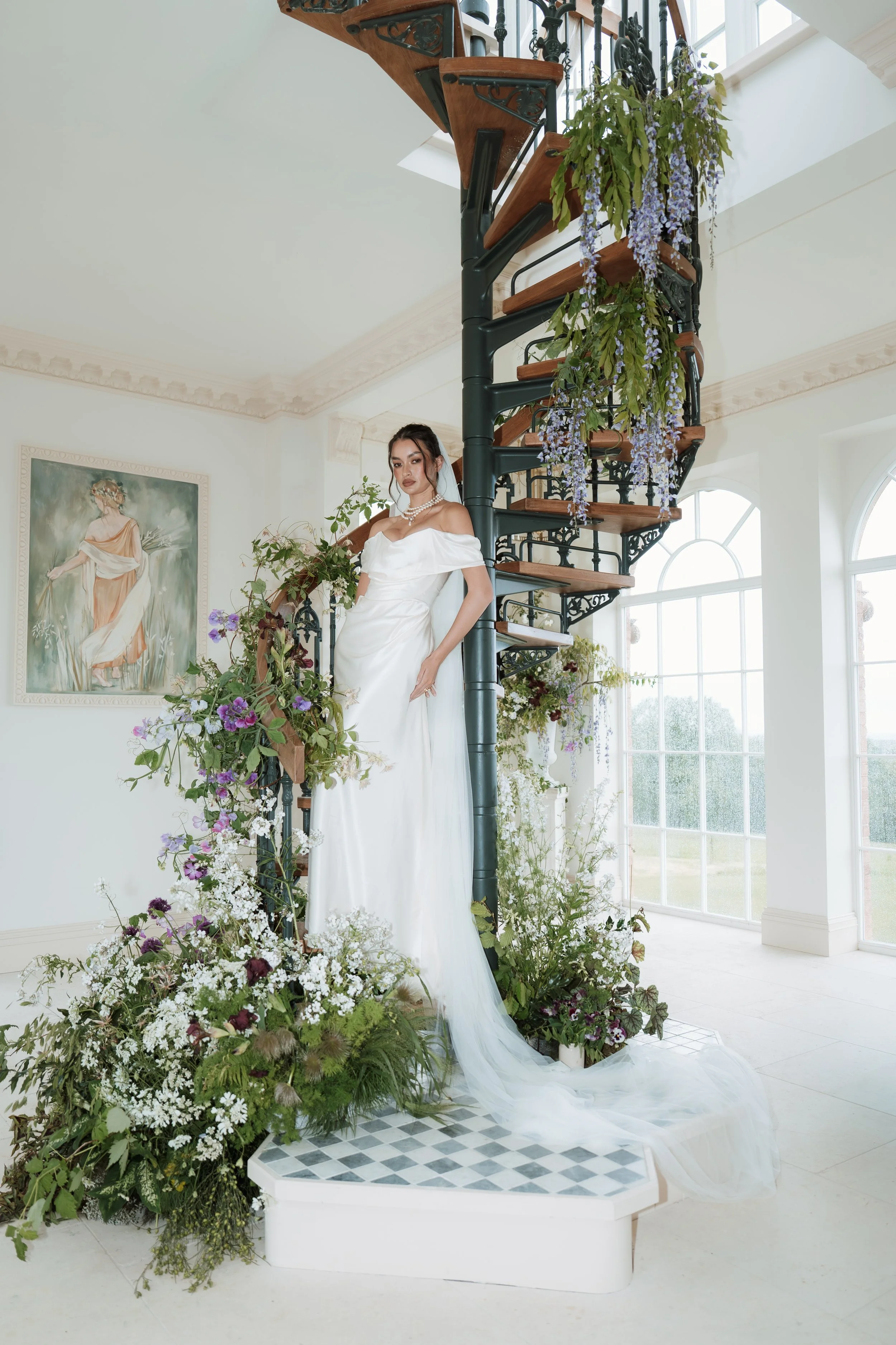 An elegant bride in a luxury Vivienne Westwood wedding dress standing on a small indoor staircase beautifully adorned with cascading white flowers and greenery.