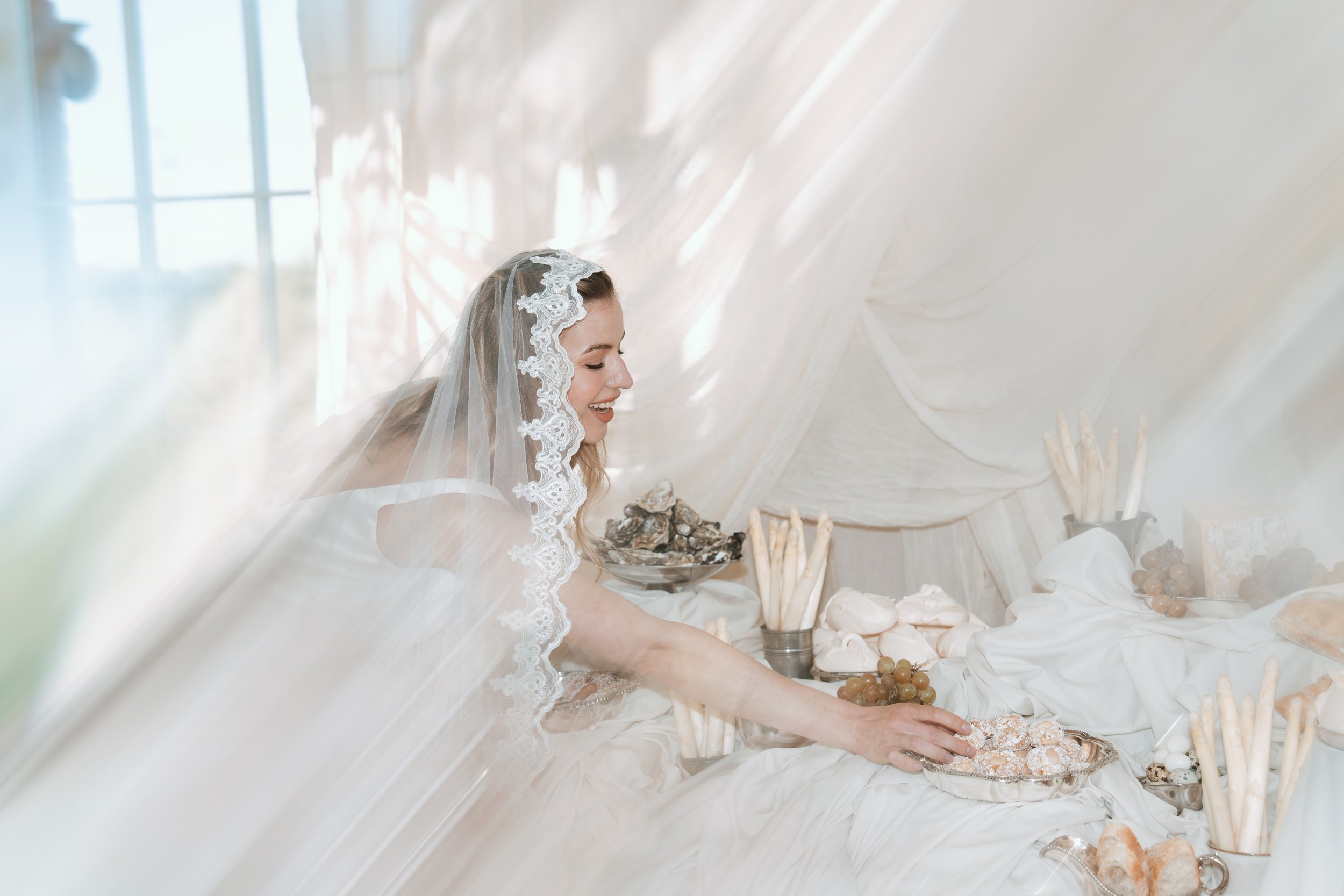 A smiling bride in an elegant Savannah Miller Bridal wedding dress and lace veil, reaching for food at a beautifully styled buffet table decorated with white fabric and seashells.