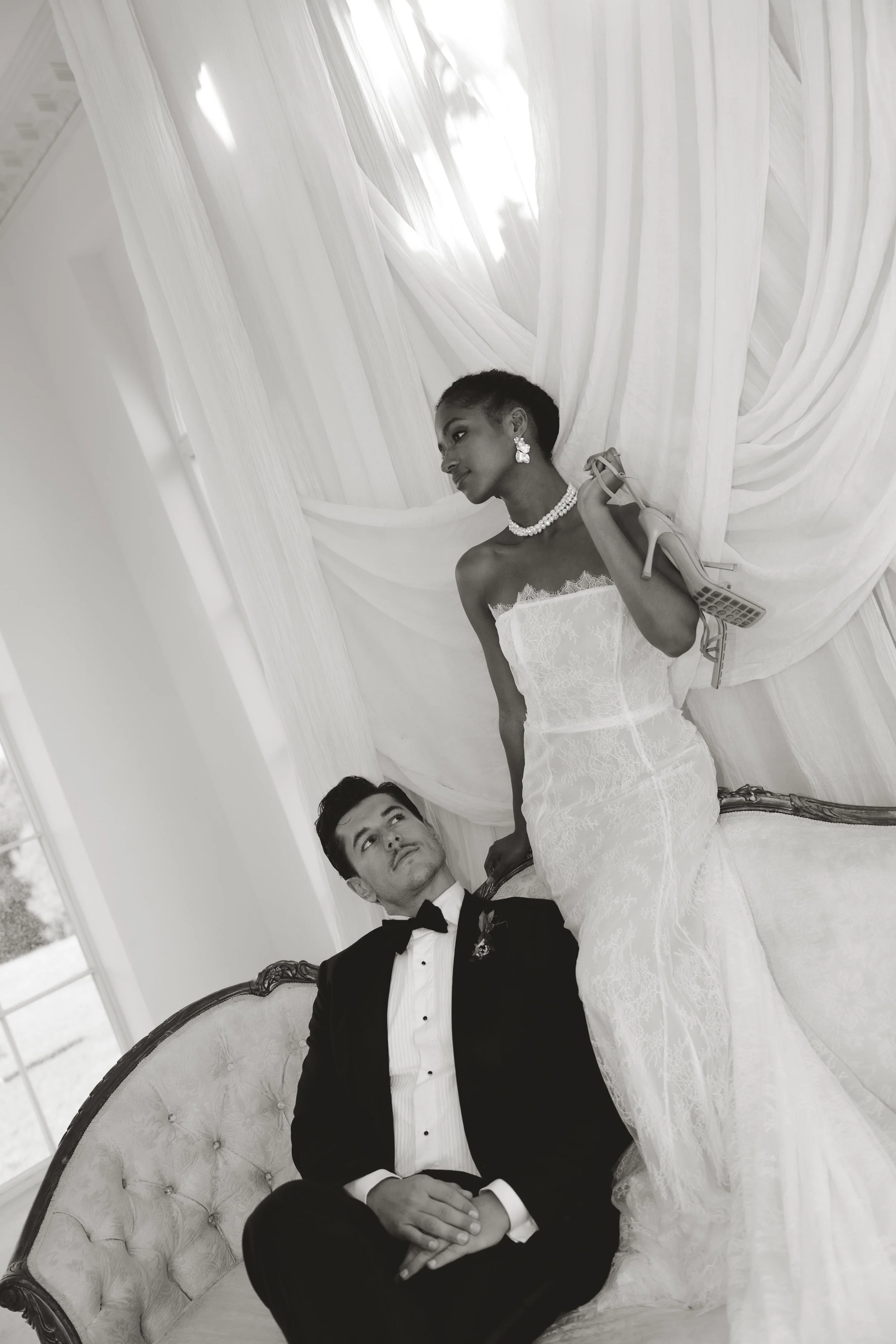 Black and white editorial bridal photography of a woman in a strapless lace wedding dress and pearl jewelry holding a high heel shoe, standing by a groom in a tuxedo on a vintage couch.
