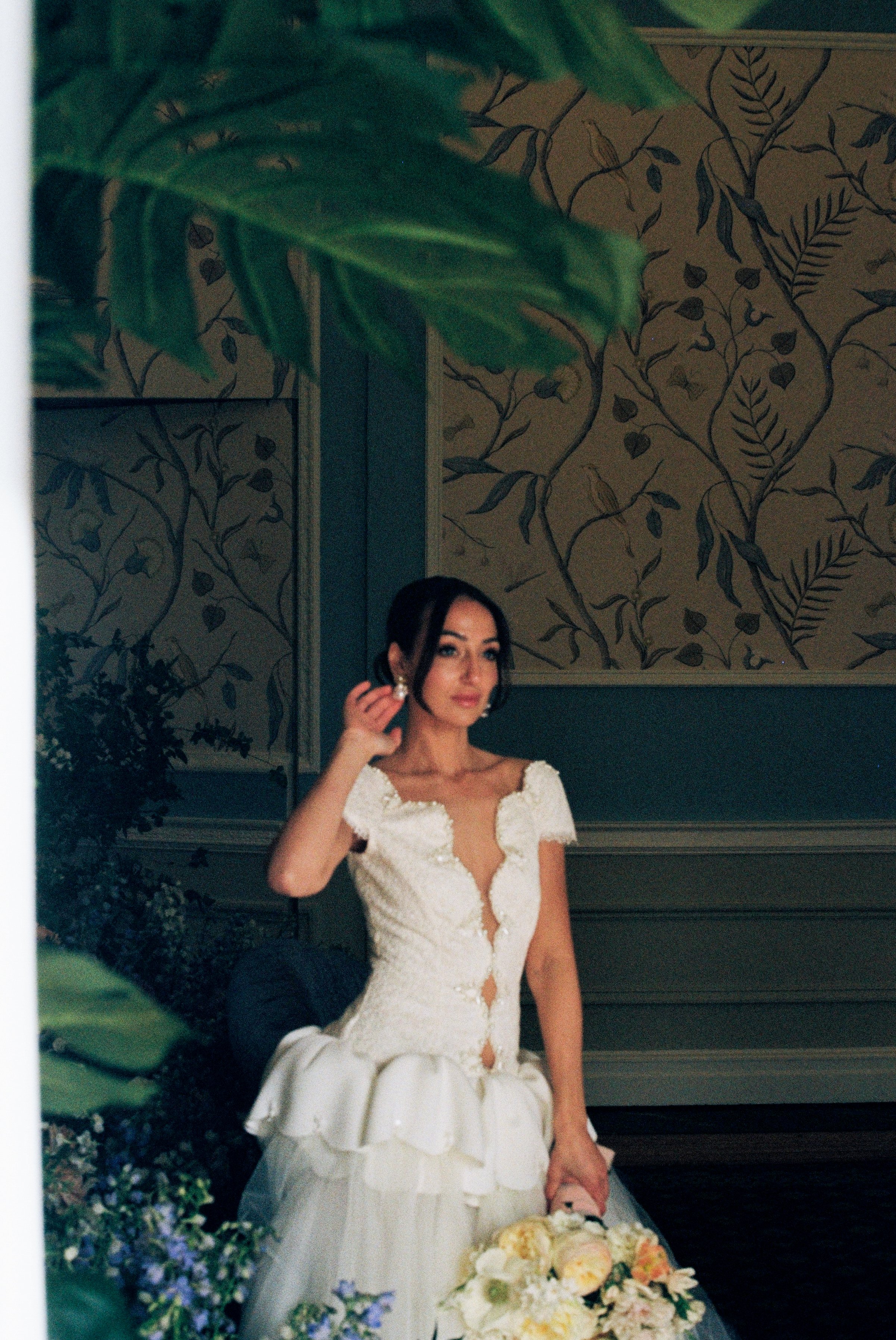A bride in a white lace wedding dress sitting with a bouquet of flowers, holding an earring, with greenery and decorated wall in the background.
