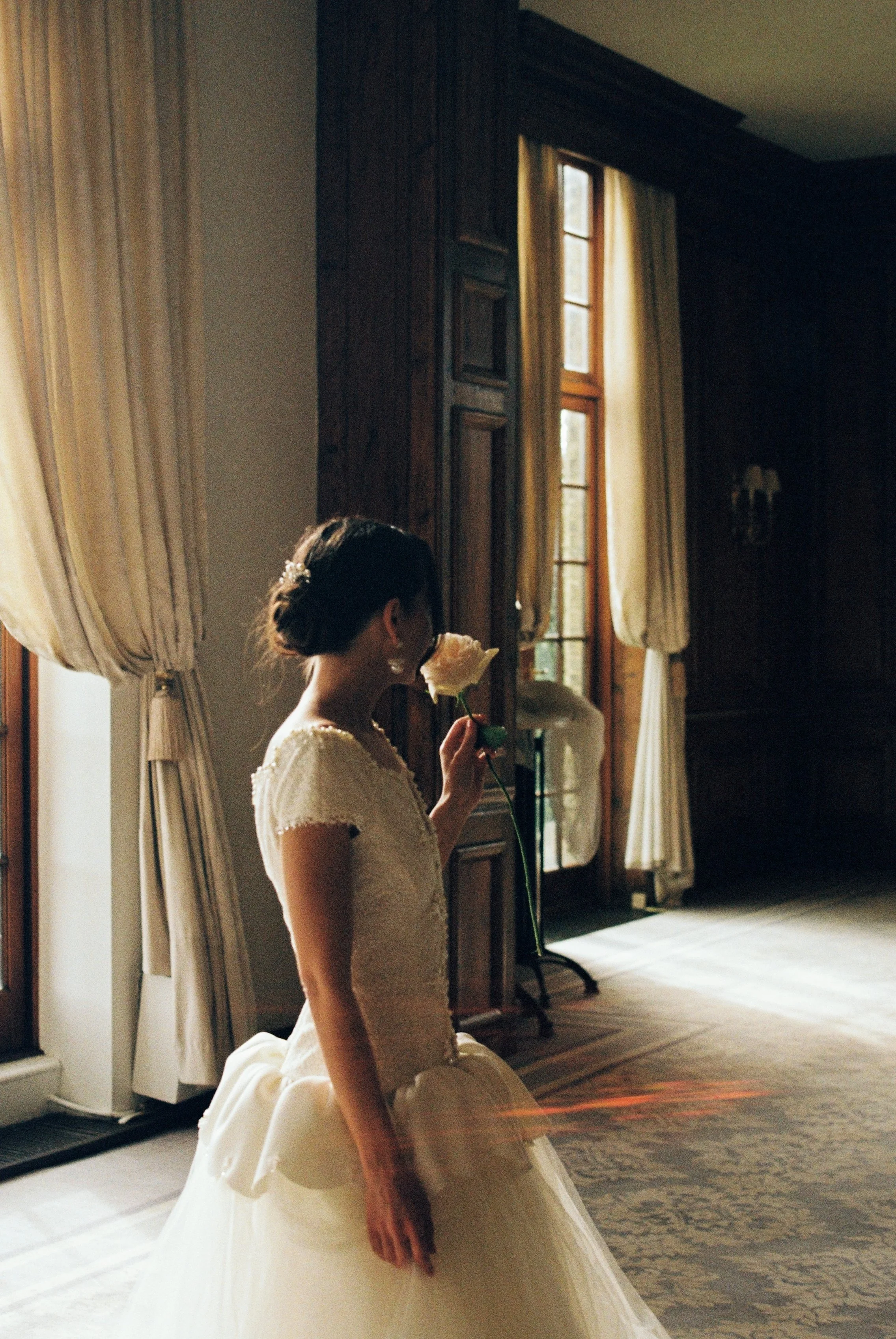 A bride in a white dress kneeling on a patterned rug, holding a single white flower, with large windows and heavy cream-colored curtains in the background.