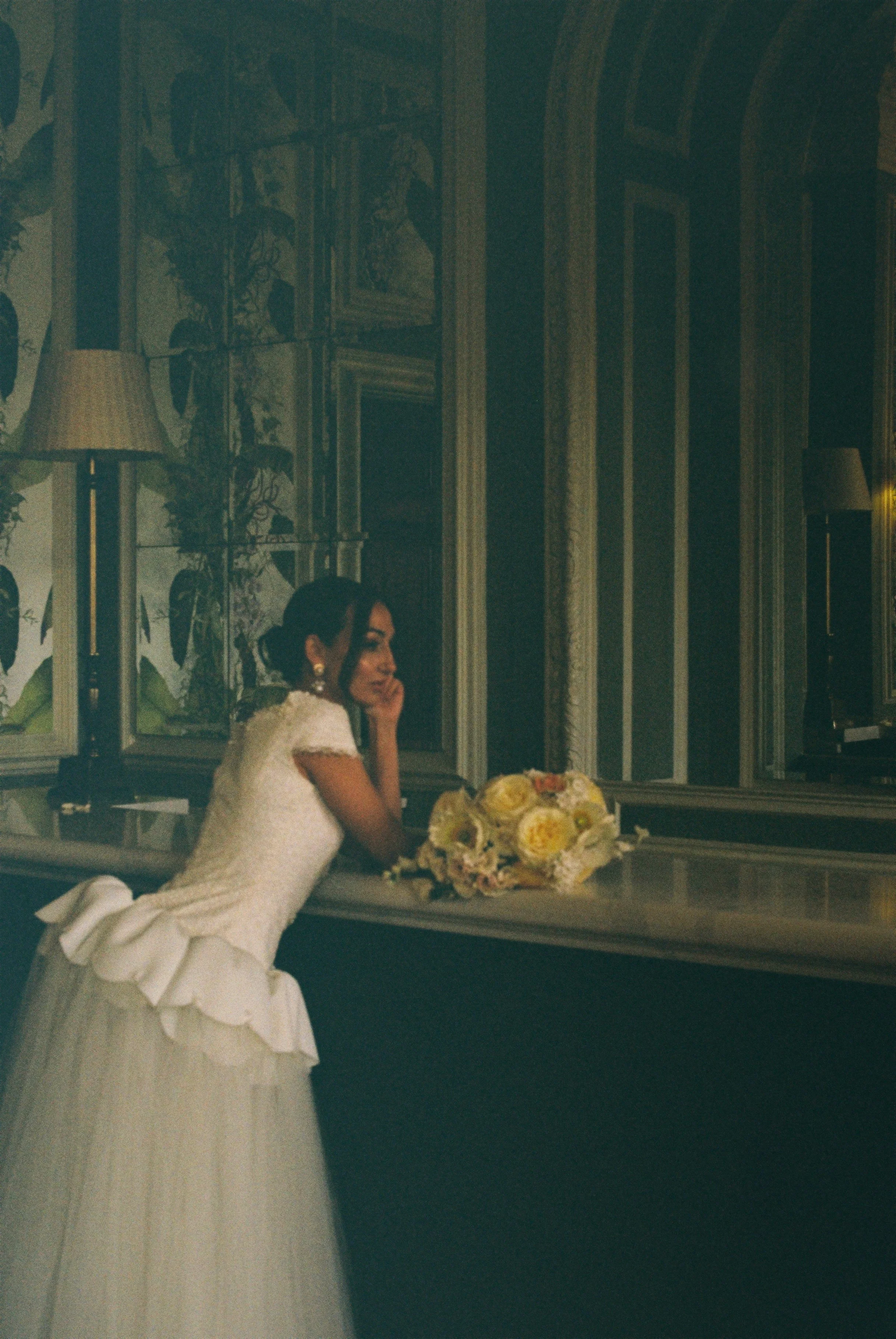 A bride in a white wedding dress sitting at a table with a bouquet of flowers, in a dimly lit room with ornate paneling and wall decorations.