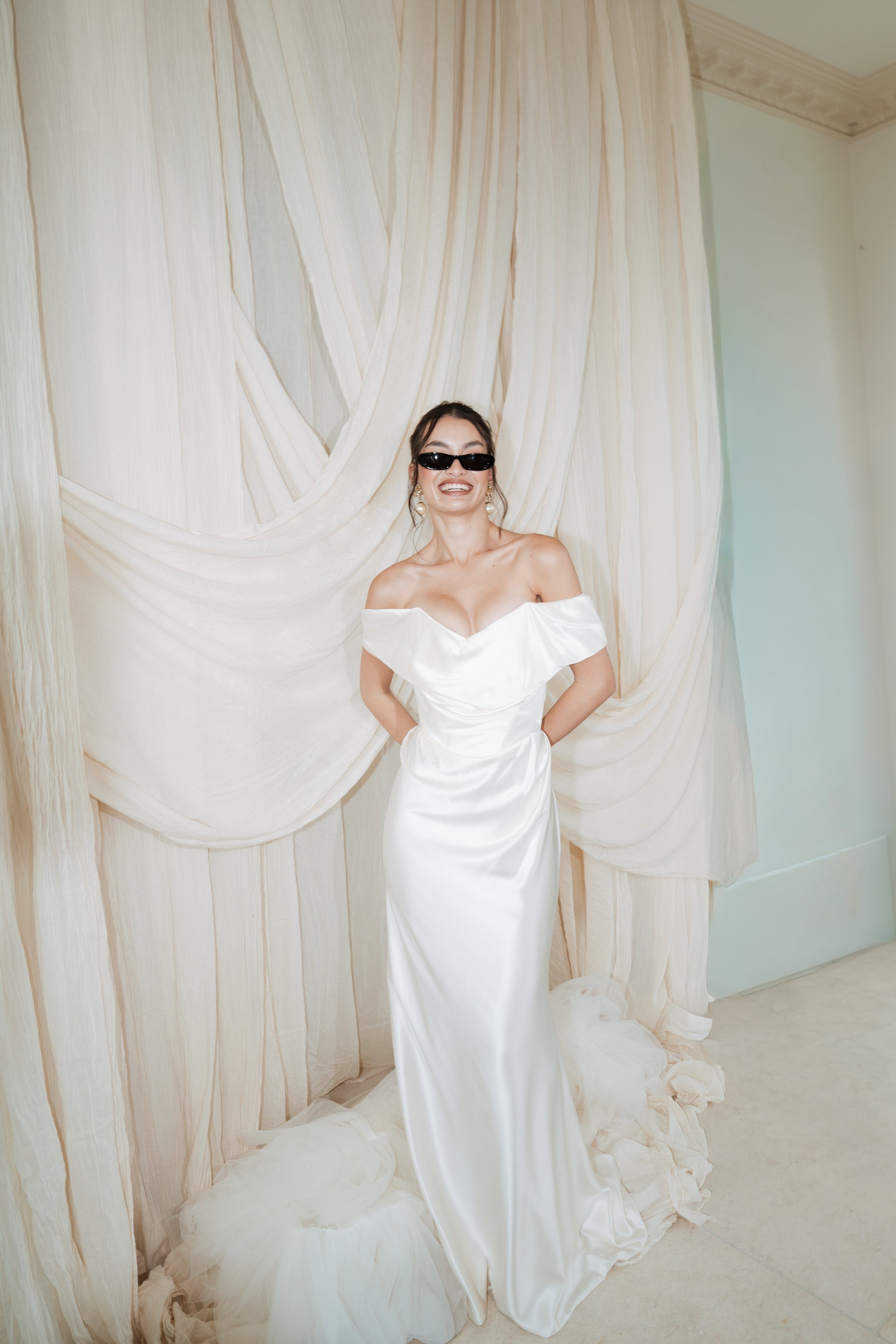 A bride wearing a white off-shoulder Vivienne Westwood dress and Saint Laurent sunglasses, smiling in front of beige draped curtains.