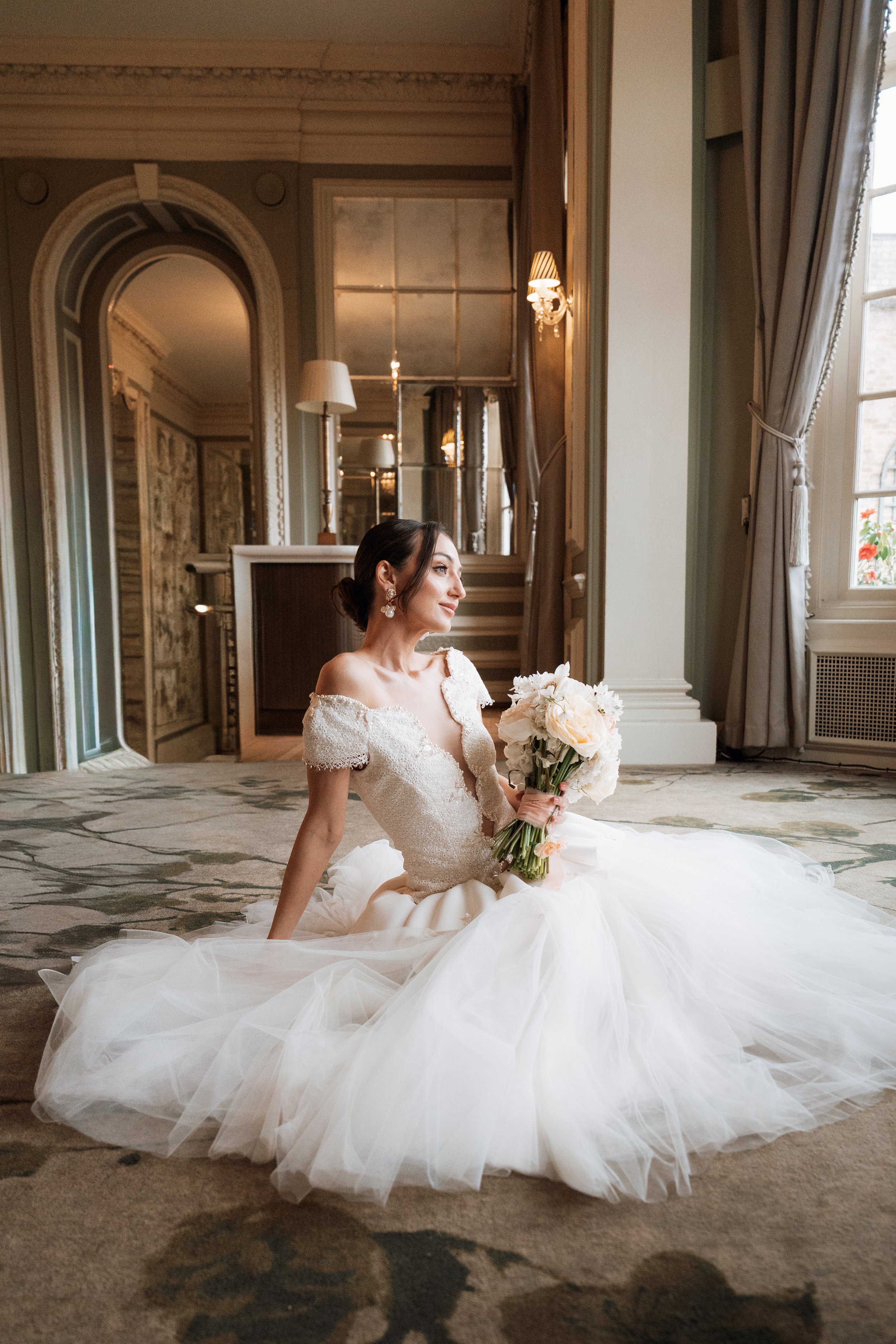 An editorial bridal portrait of a woman in a voluminous white wedding gown sitting on the floor of a grand, luxurious room at Hedsor House, holding a white bouquet.