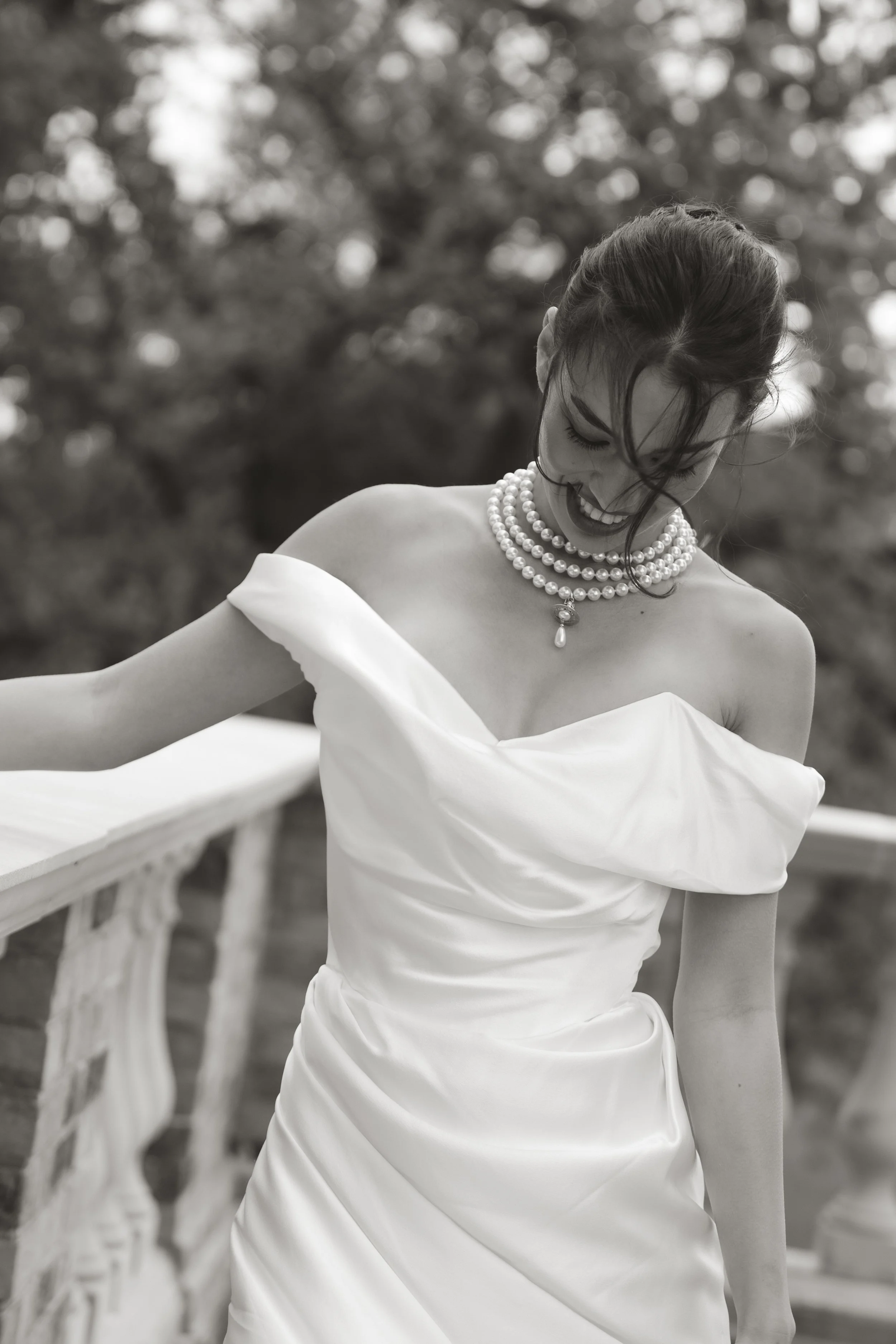 Black and white wedding portrait of an elegant bride wearing a strapless Vivienne Westwood wedding dress and multiple pearl necklaces, looking down and smiling outdoors.