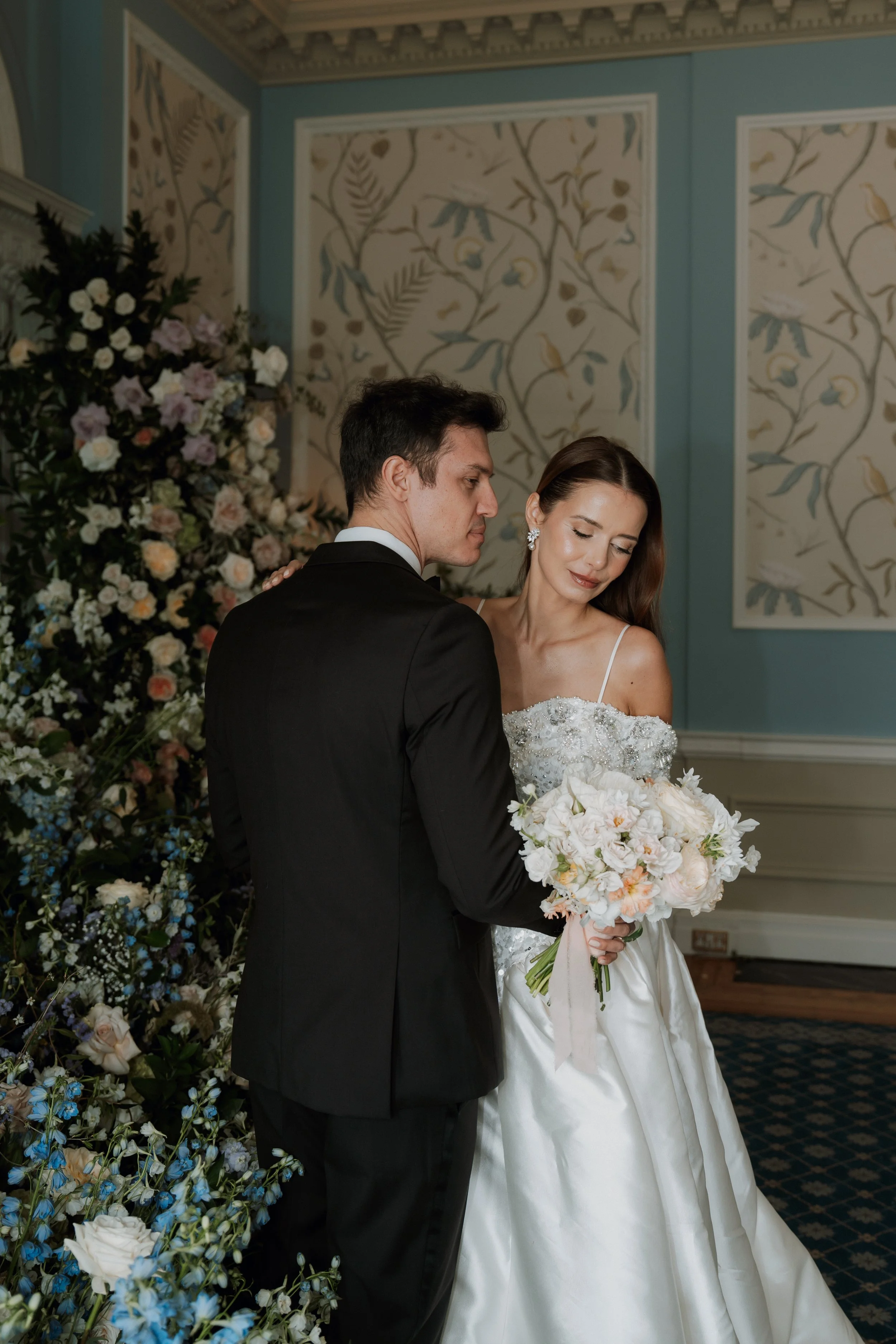 An elegant bride and groom posing indoors at Hedsor House, surrounded by exquisite floral installations and stately wallpapered walls.