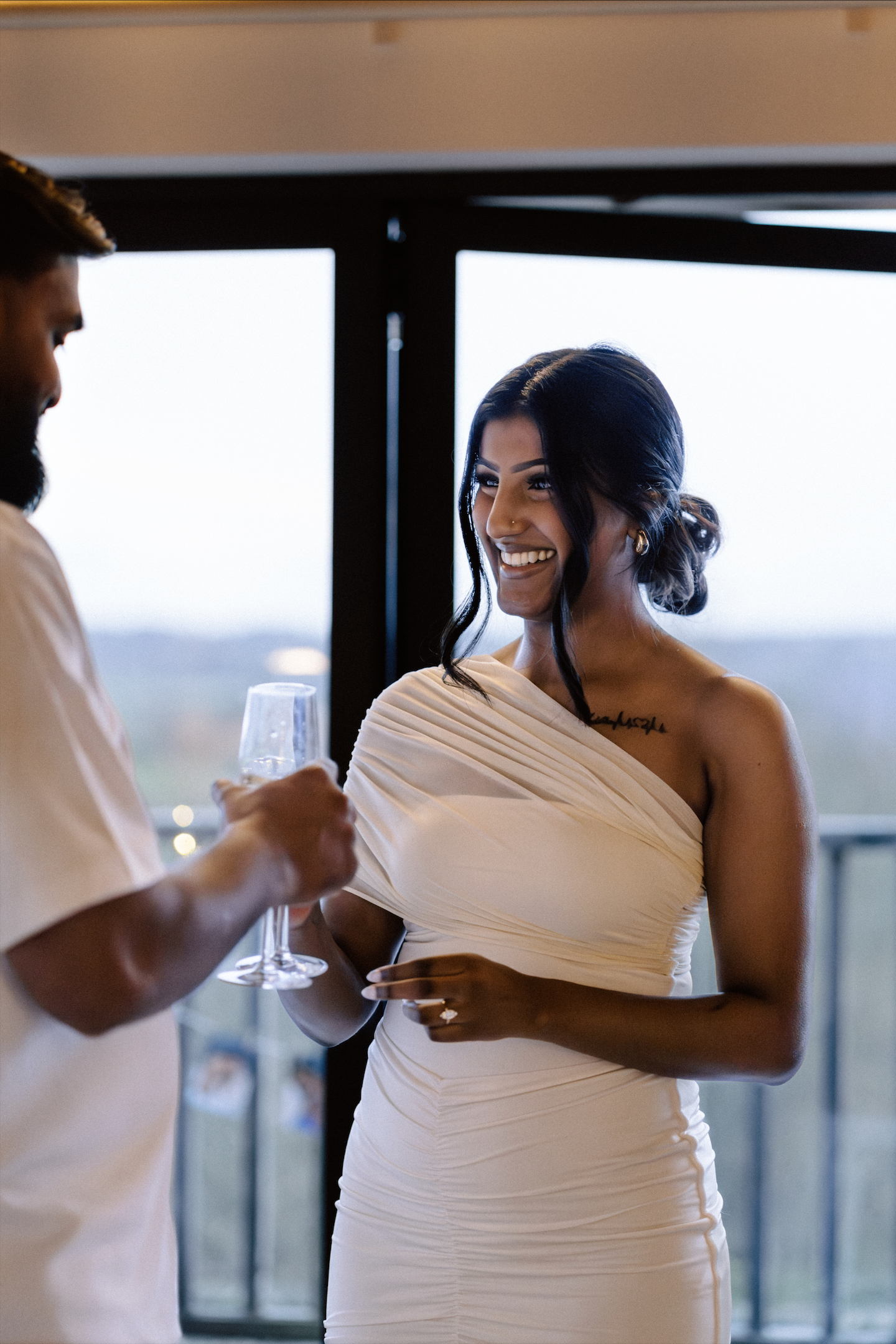 An elegant bride in a luxury white wedding dress smiling as she receives a drink at a chic, contemporary wedding reception.