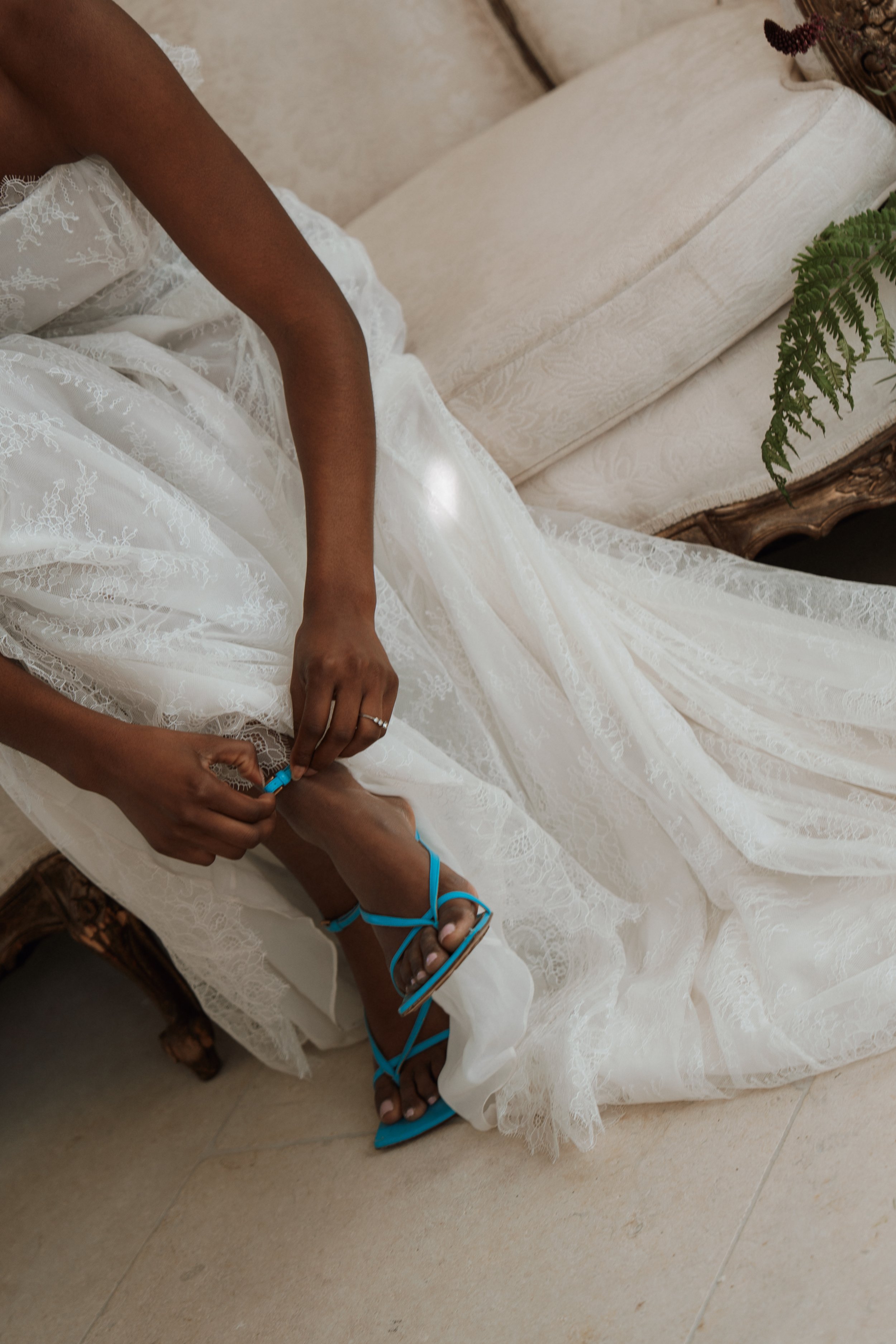 Close-up bridal detail shot of a woman in a luxury Vera Wang lace wedding dress sitting on a beige couch, adjusting a statement bright blue high heel sandal on her foot.