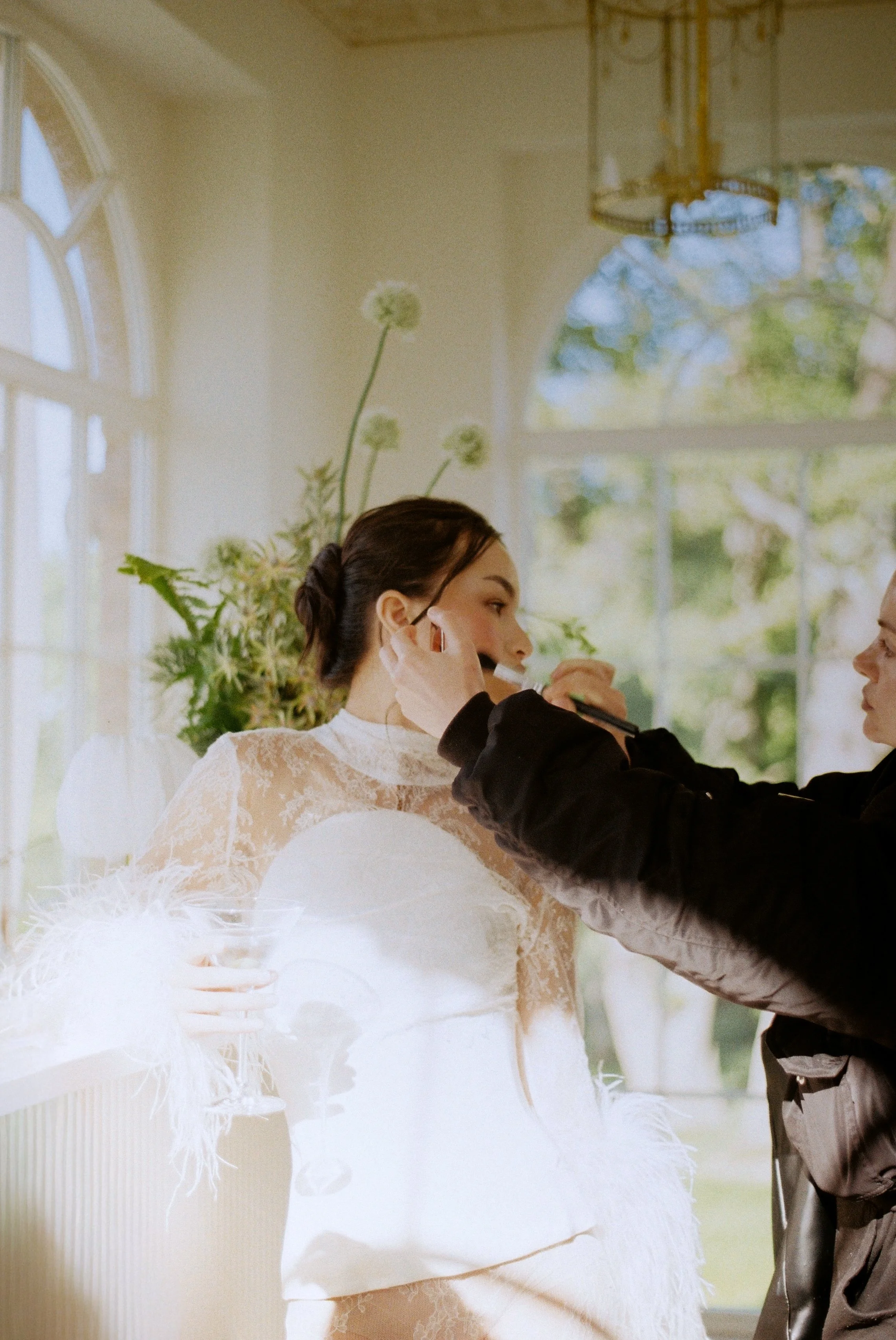 A Bride in a white lace dress gets ready for her wedding, with the makeup artist nearby, in a bright room with large windows and green plants.