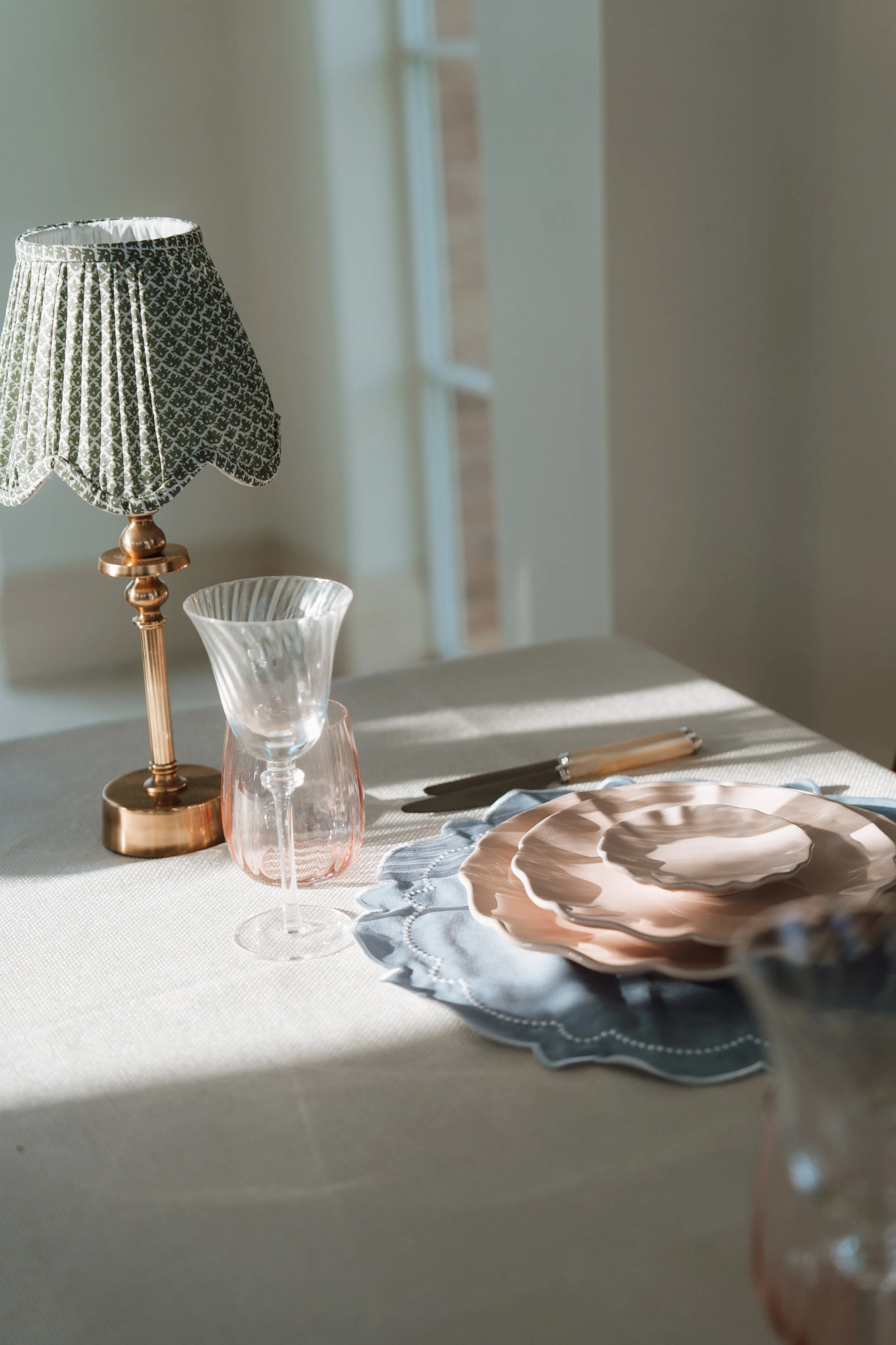 A table setting with a vintage-style table lamp with a black and white patterned fabric shade, pink-tinted glassware, a place setting with stacked pink and white plates, and a silver spoon. Sunlight streams through a window in the background.