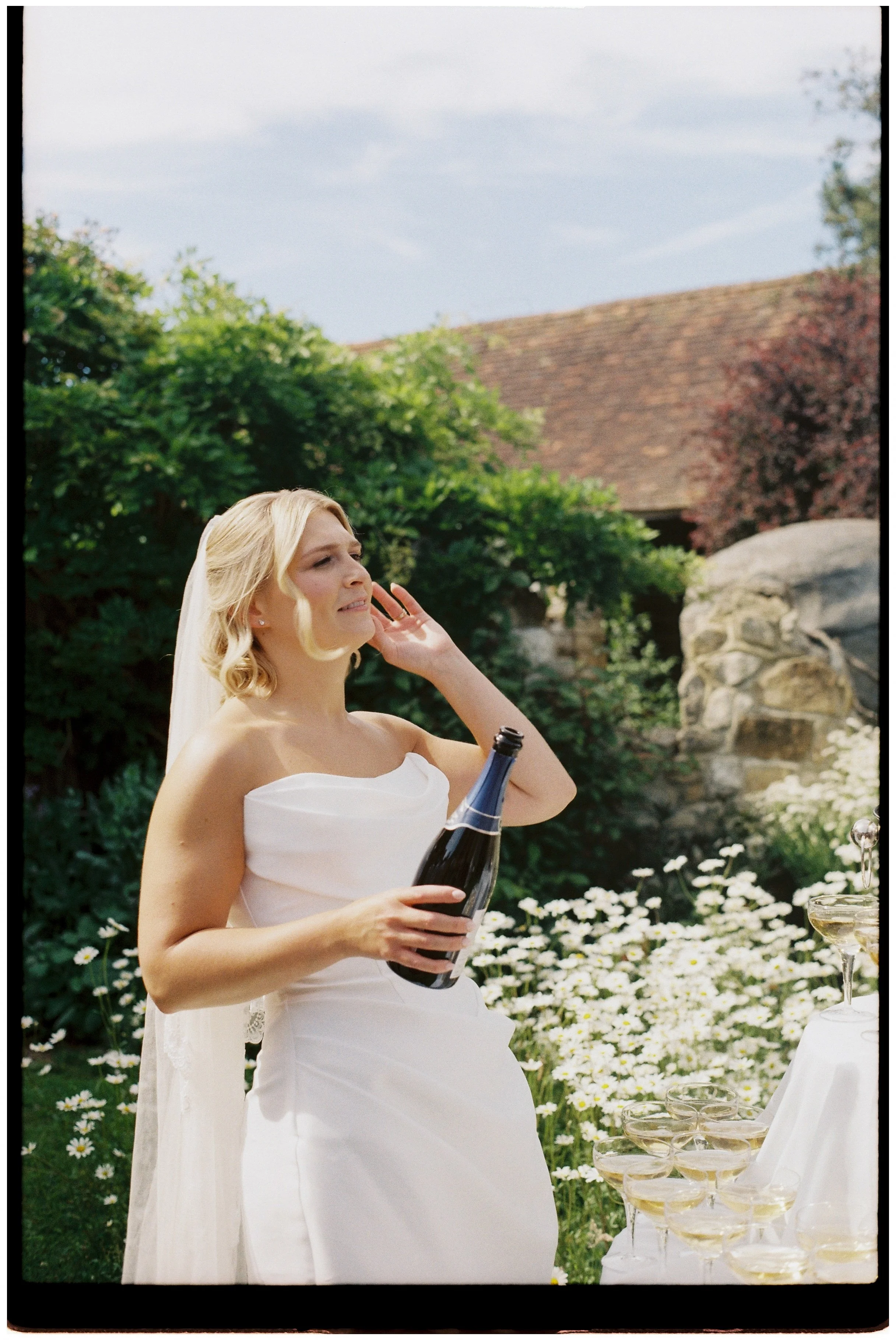 A bride in a white wedding dress and veil at an outdoor celebration, holding a champagne bottle, with a table of champagne glasses and white flowers nearby, and greenery and a stone building in the background.