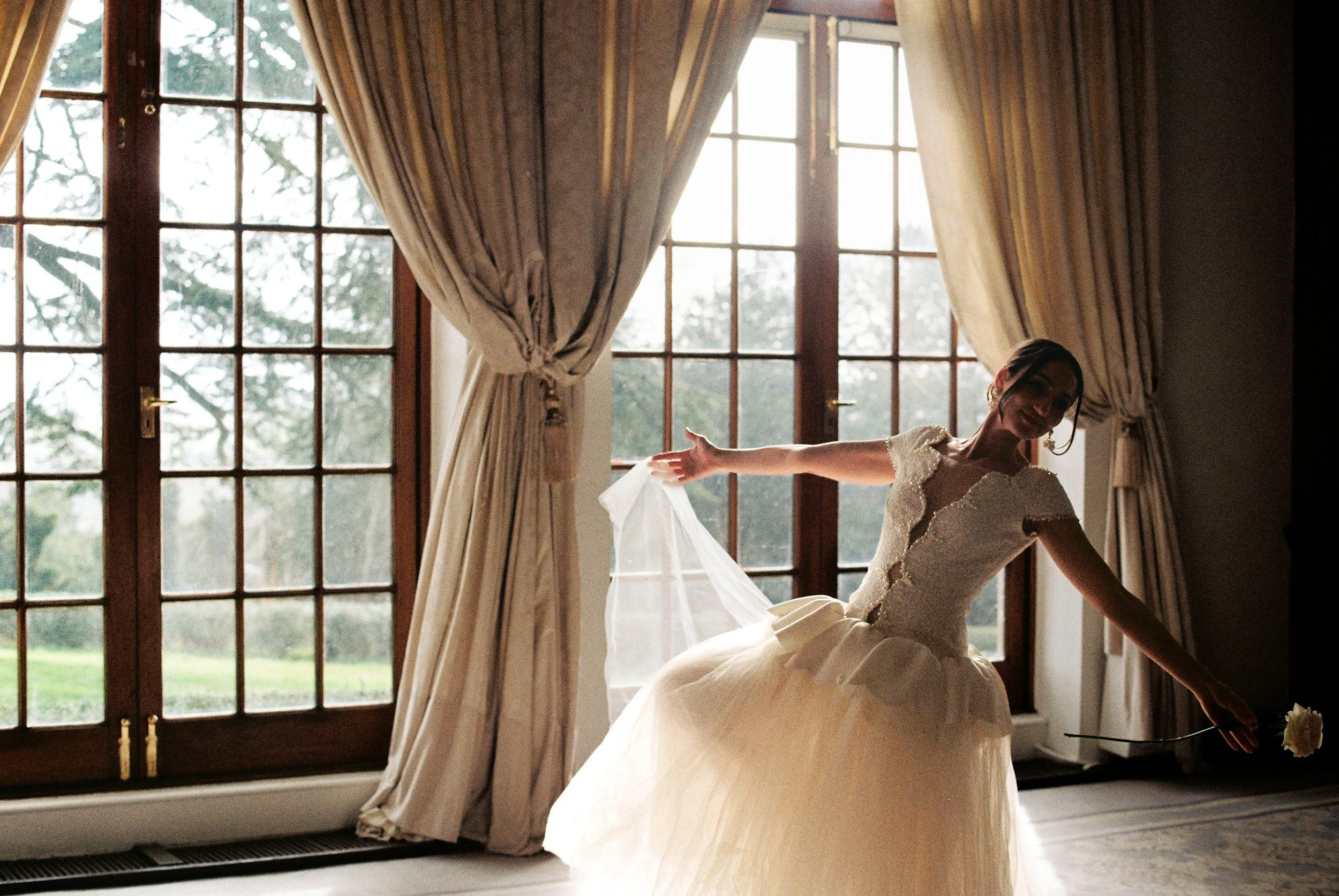 A bride in a wedding dress standing near large windows at Hedsor House, smiling and holding her dress with one hand in a warmly lit room.