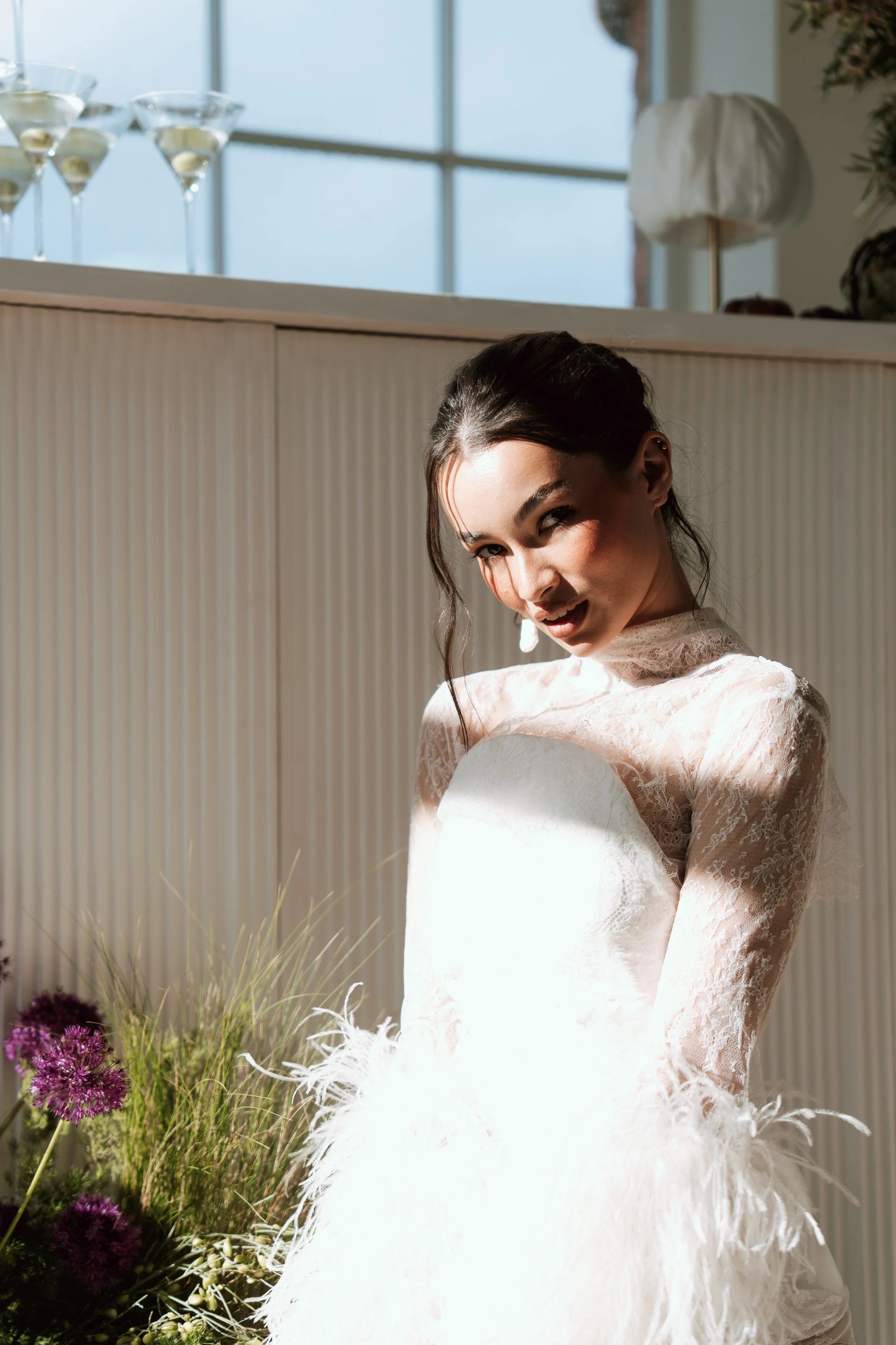 An elegant bride wearing a luxury Asar London feather wedding dress with white lace details, holding a bouquet and standing indoors near a window with a beautiful floral arrangement.