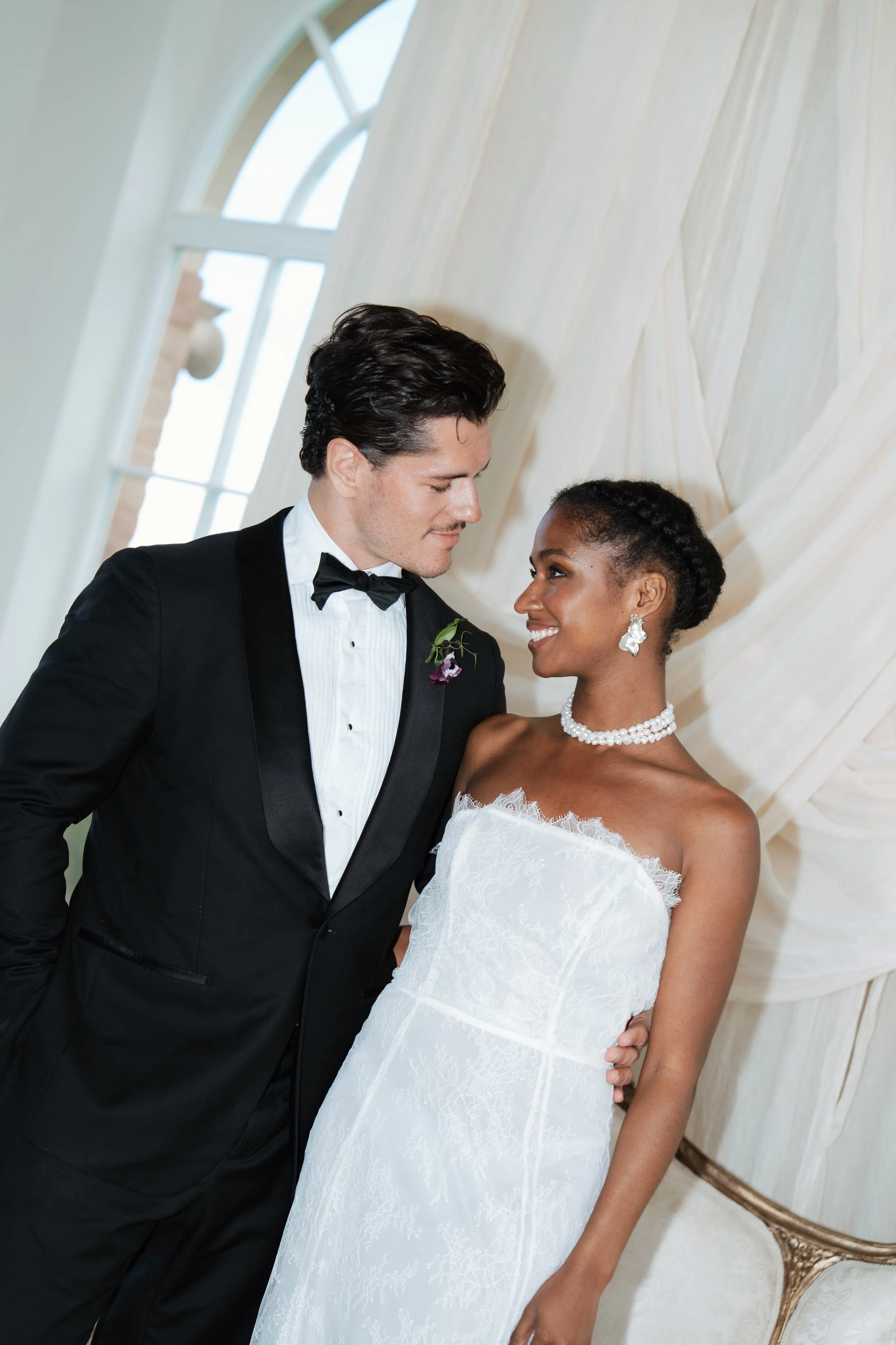 A romantic indoor portrait of a bride in a luxury white wedding dress and a groom in a classic black tuxedo, standing close together in a beautifully lit room.