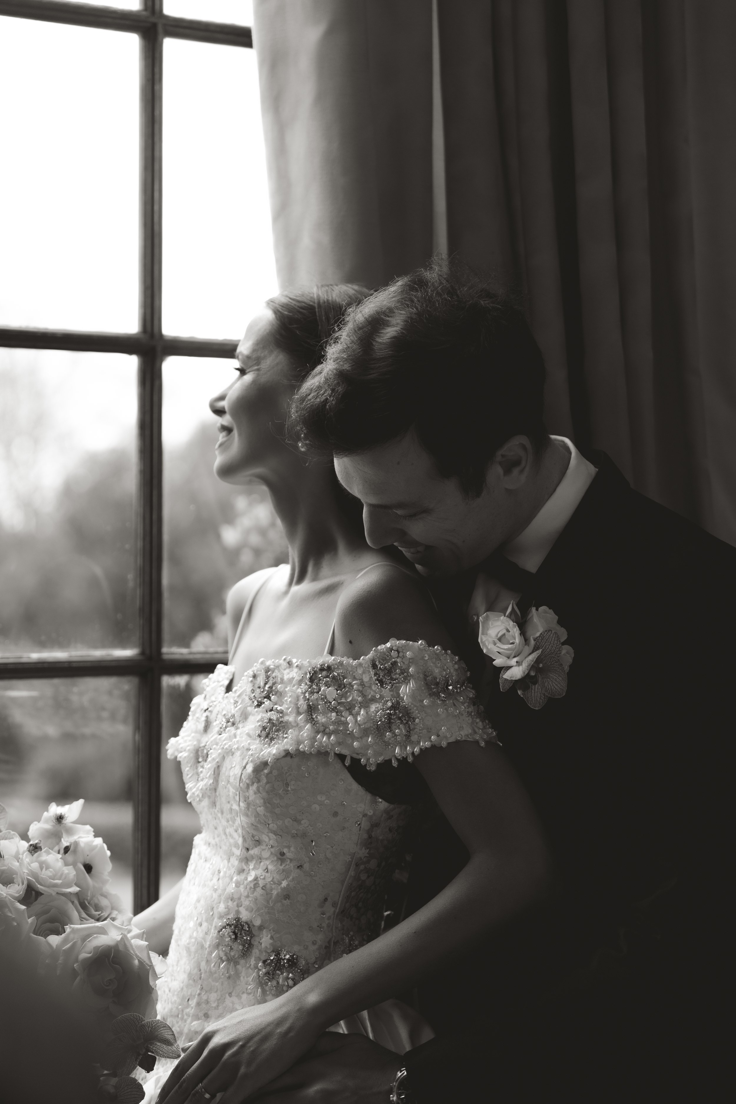 Black and white editorial wedding photography of a bride and groom leaning close together, sharing an intimate smile next to a window at Hedsor House.