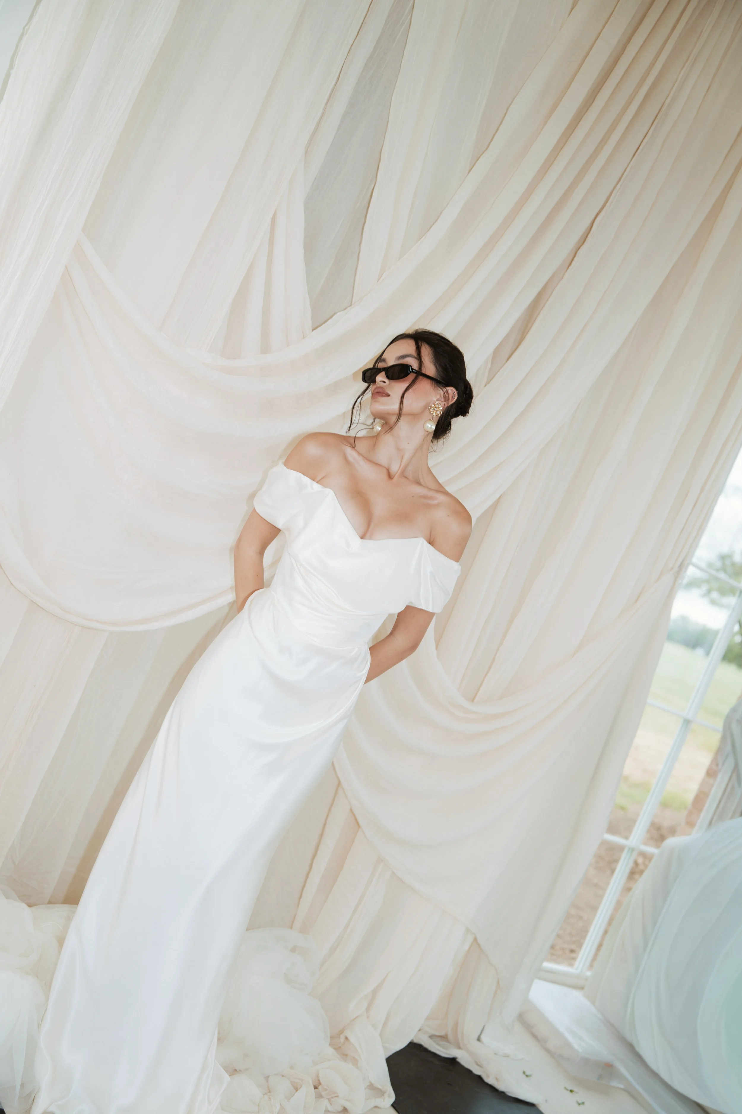 Chic and modern bridal photography of a woman styling a Vivienne Westwood wedding dress with statement Saint Laurent black sunglasses and earrings, posing against cream draped fabric.