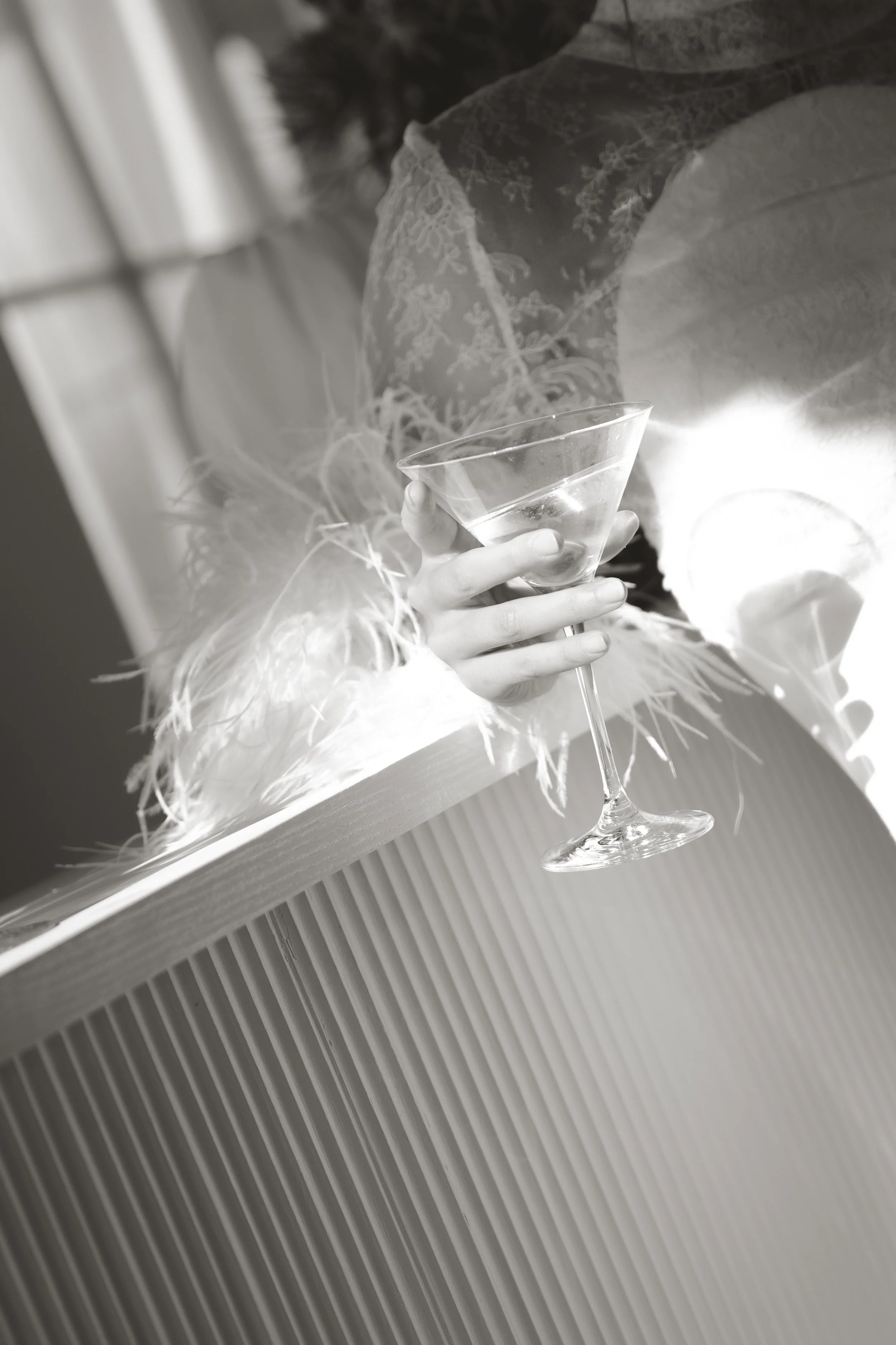 A bride celebrating with a martini glass filled, with a backdrop of a ornate curtain and decorative feathers.