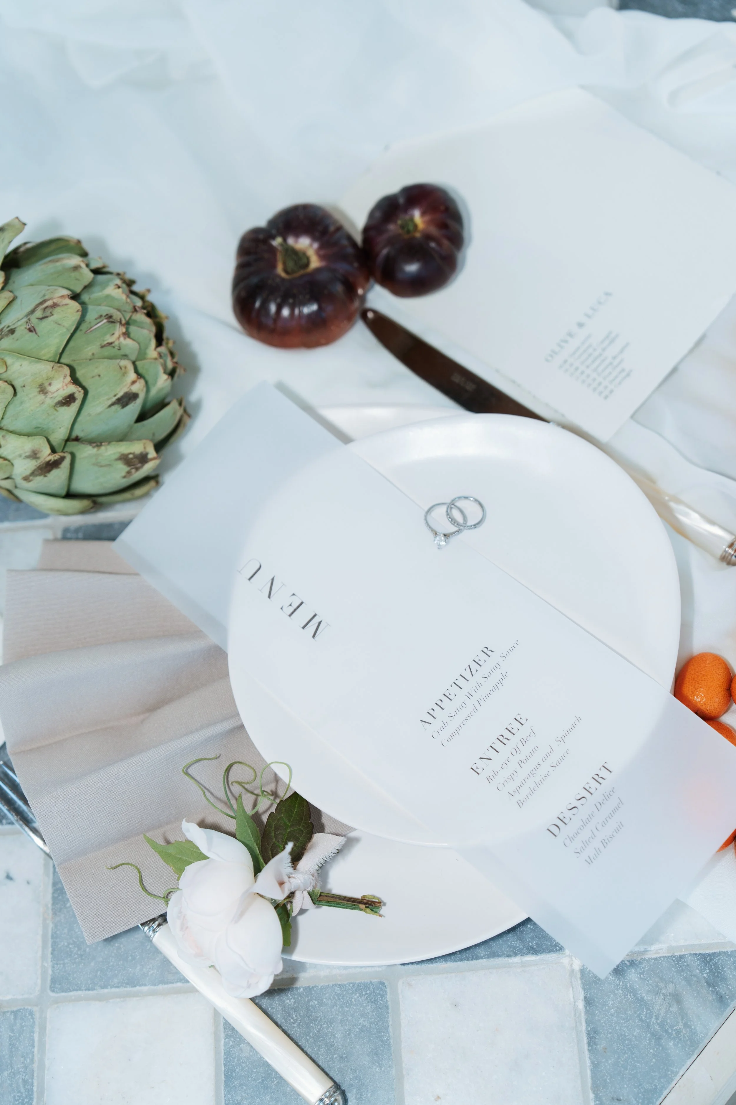 A luxury wedding flat lay featuring an elegant menu card on a white plate, paired with silver wedding rings, delicate florals, and a unique green artichoke accent on a tiled surface.