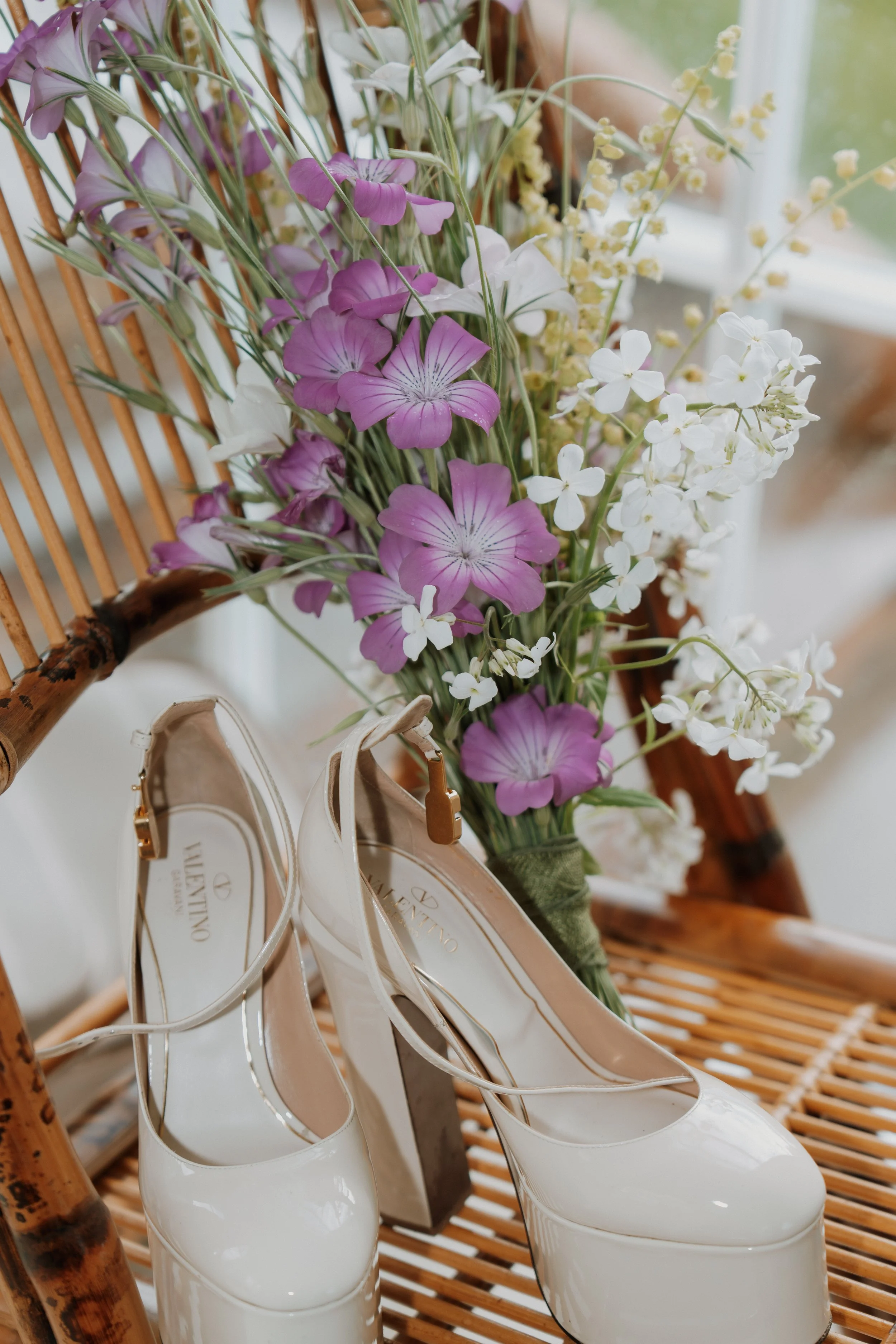 A bouquet of purple and white flowers, including lilies and other small blooms, placed on a wooden chair with white Valentino shoes hanging from the chair arms.