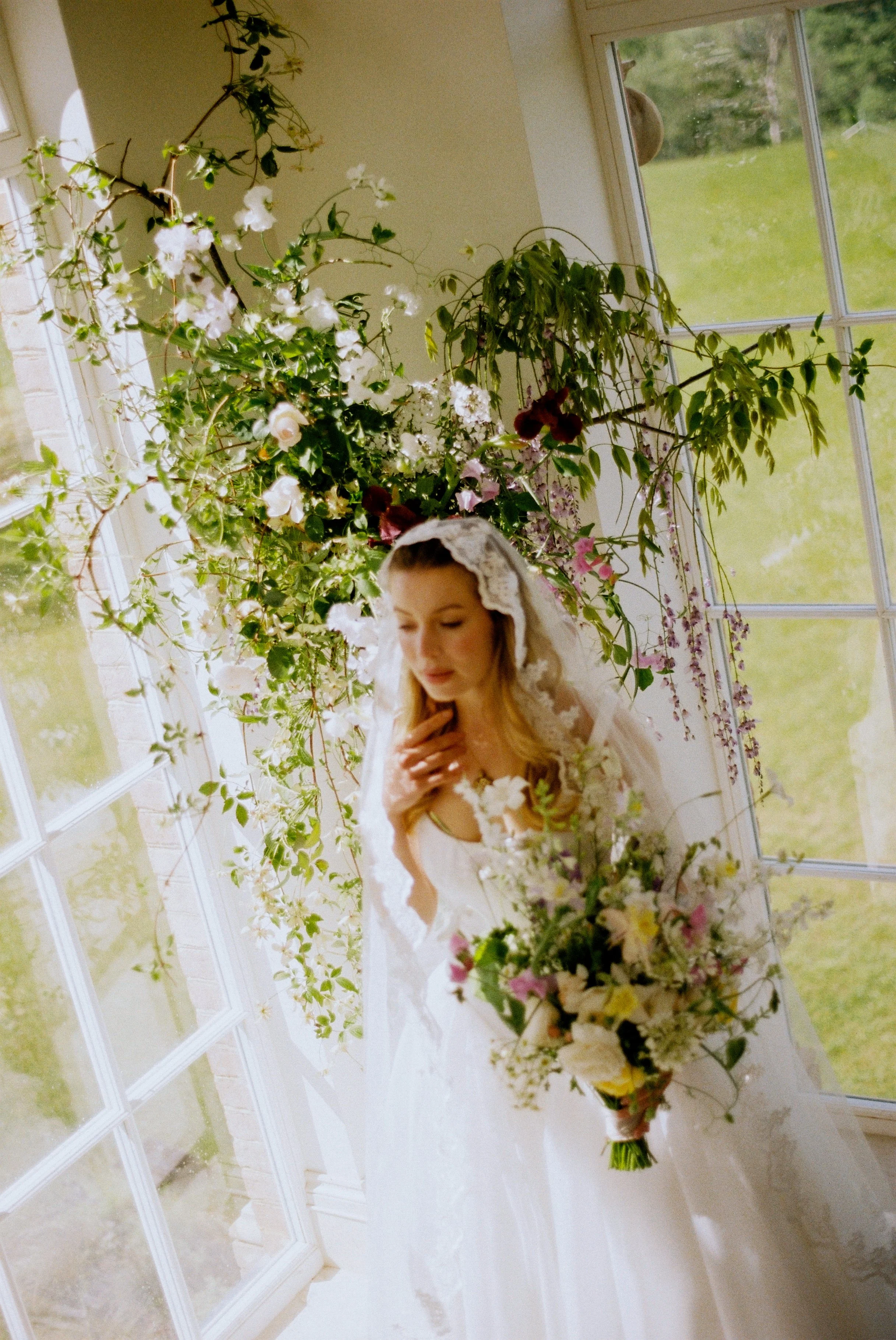 A bride in a white wedding dress and veil is standing indoors near a large window with a green lawn outside. She is holding a bouquet of flowers and appears to be smiling or looking down.