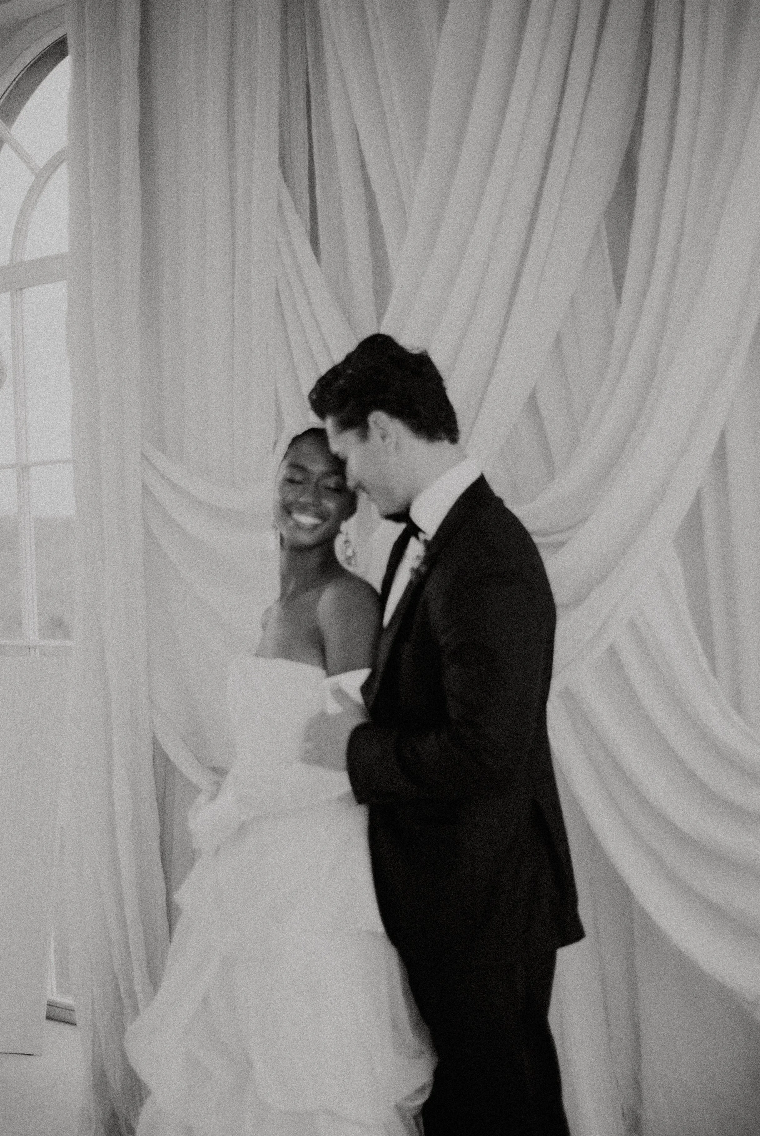 A smiling couple dressed in wedding attire, standing close together indoors in front of a decorative draped curtain. The bride is wearing a strapless wedding gown and the groom is in a tuxedo, with their foreheads touching.