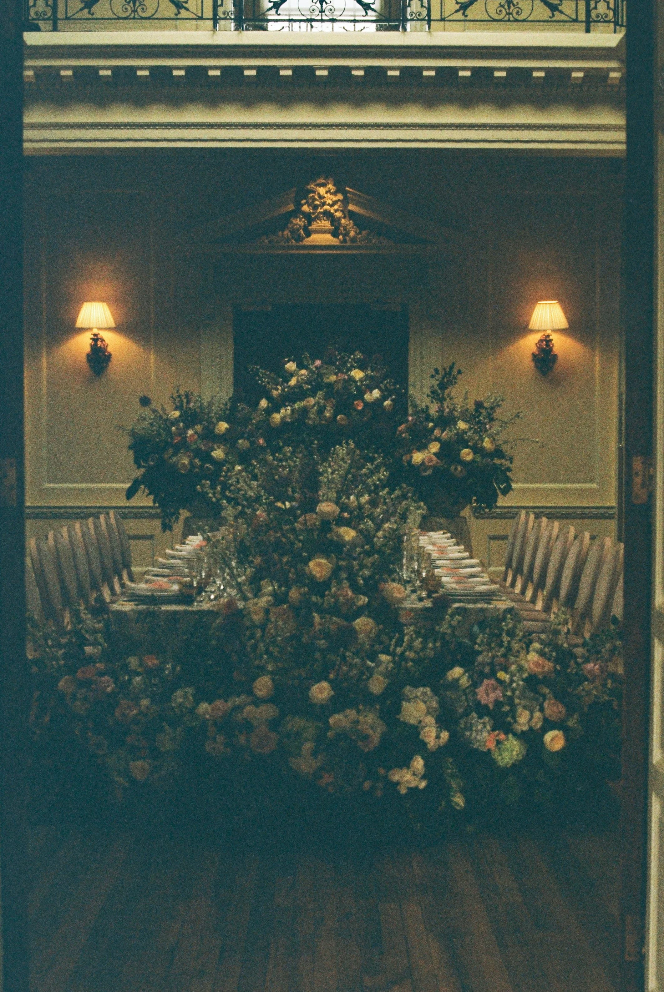 A long dining table with a large floral centerpiece, set with plates and silverware, in a formal, dimly lit room with wall sconces and a decorative ceiling. There is a balcony with ornate railing at the top of the image.