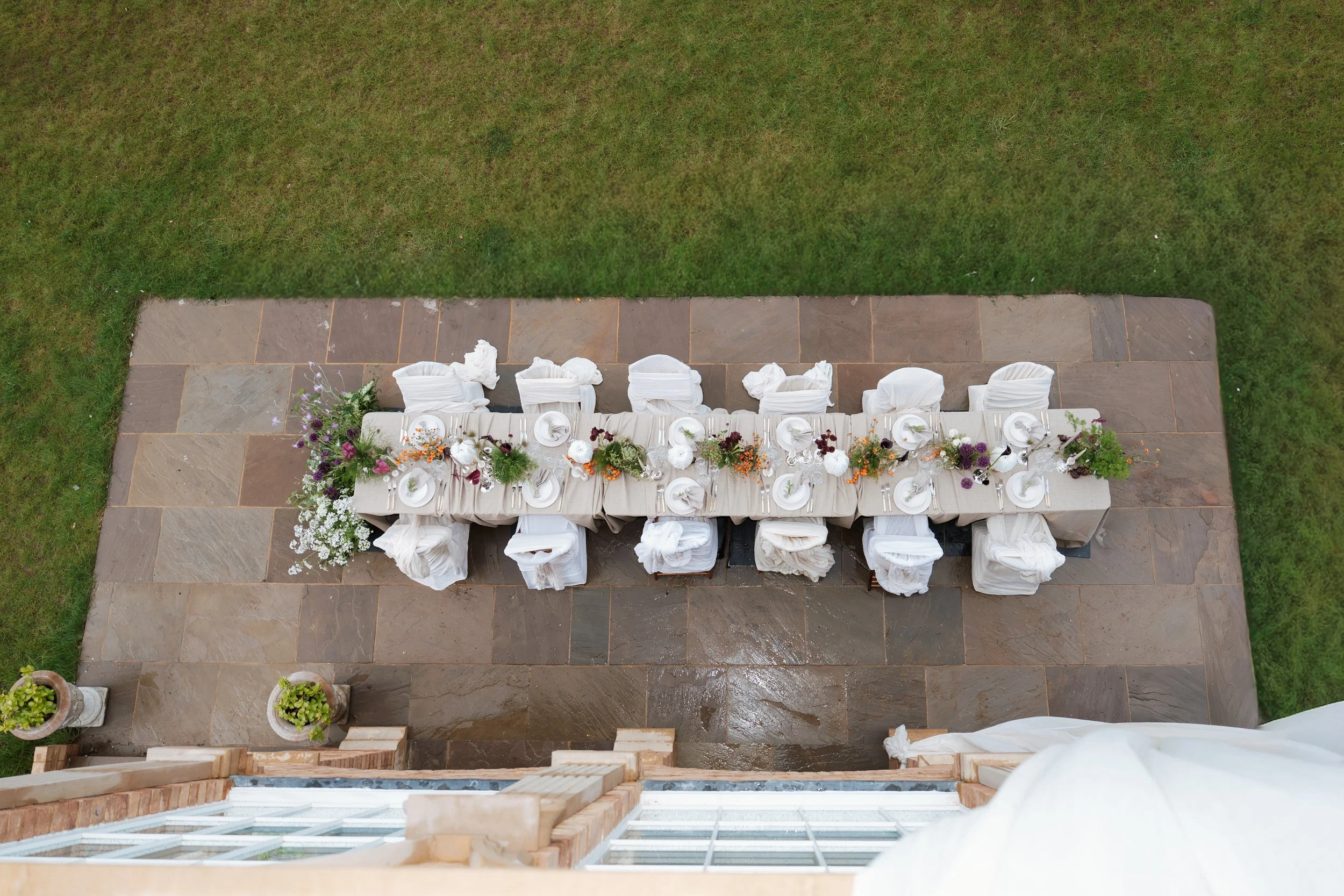 Overhead view of a long banquet table set with white cloths, plates, glasses, silverware, and floral arrangements, on a stone patio with grass surrounding it.