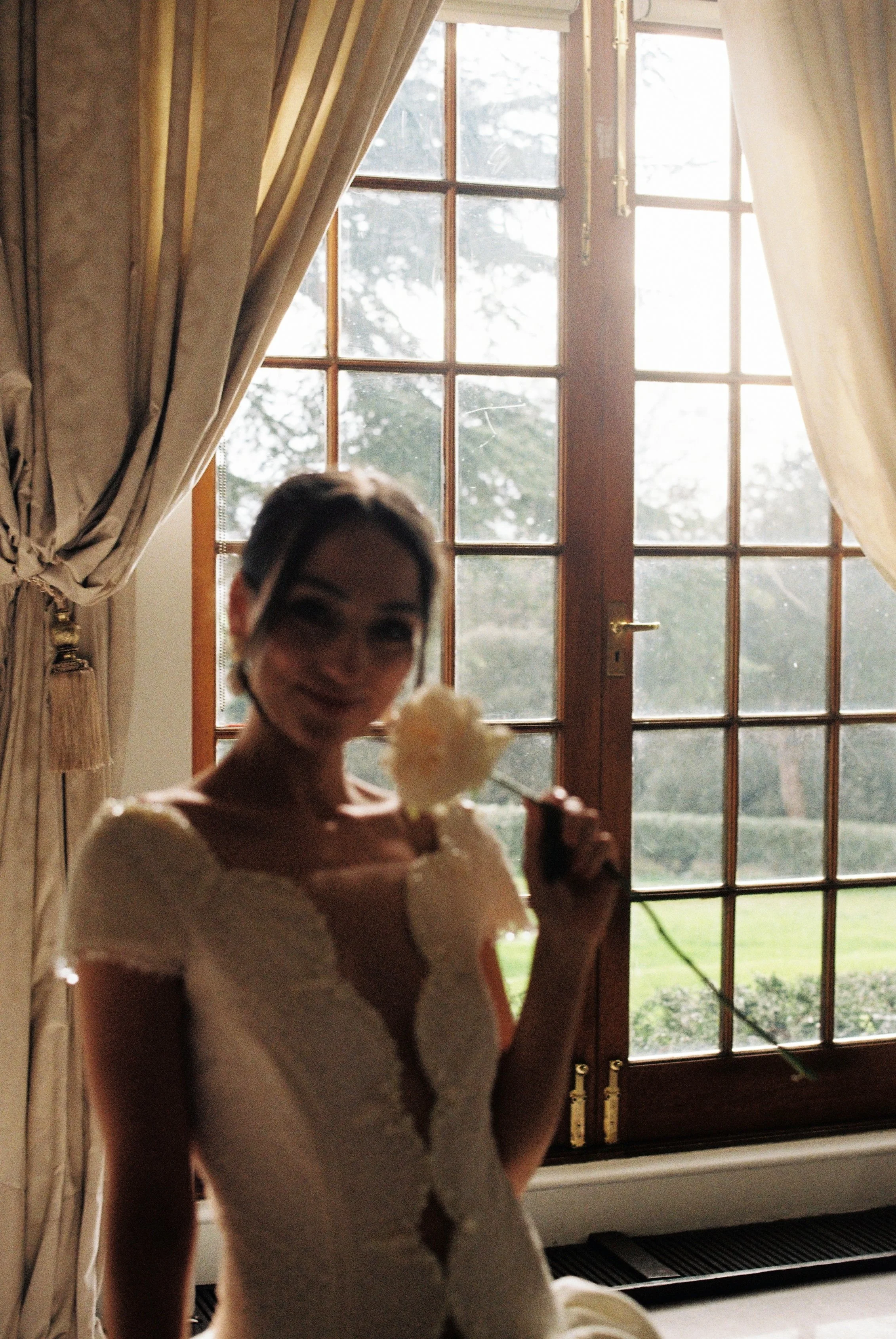 A bride in a white dress holding a light-colored flower in front of a glass door with curtains on either side, outdoors with greenery visible through the glass.