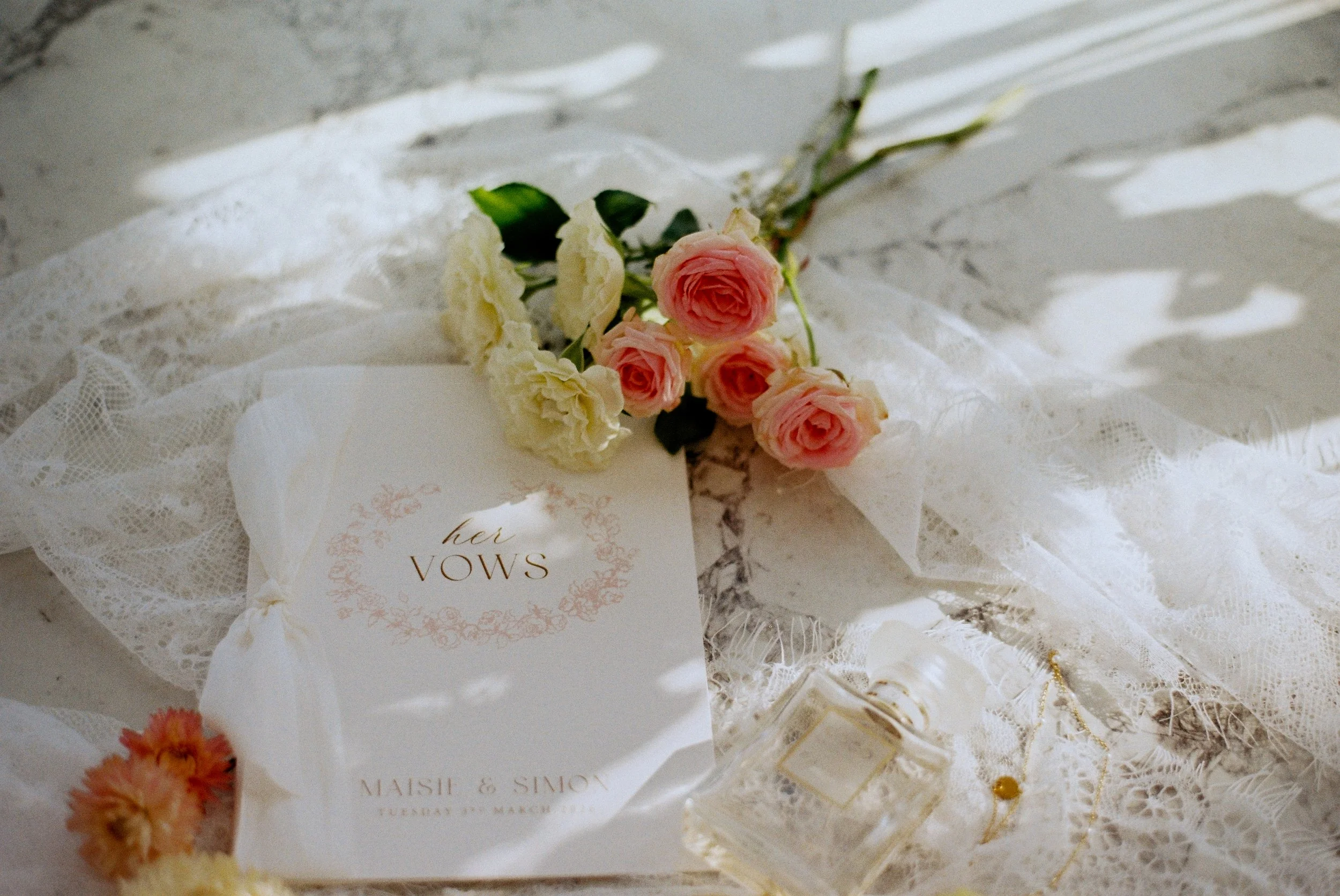 A bouquet of pink and white flowers resting on lace fabric next to a wedding invitation card that reads "her VOWS" in elegant script. 