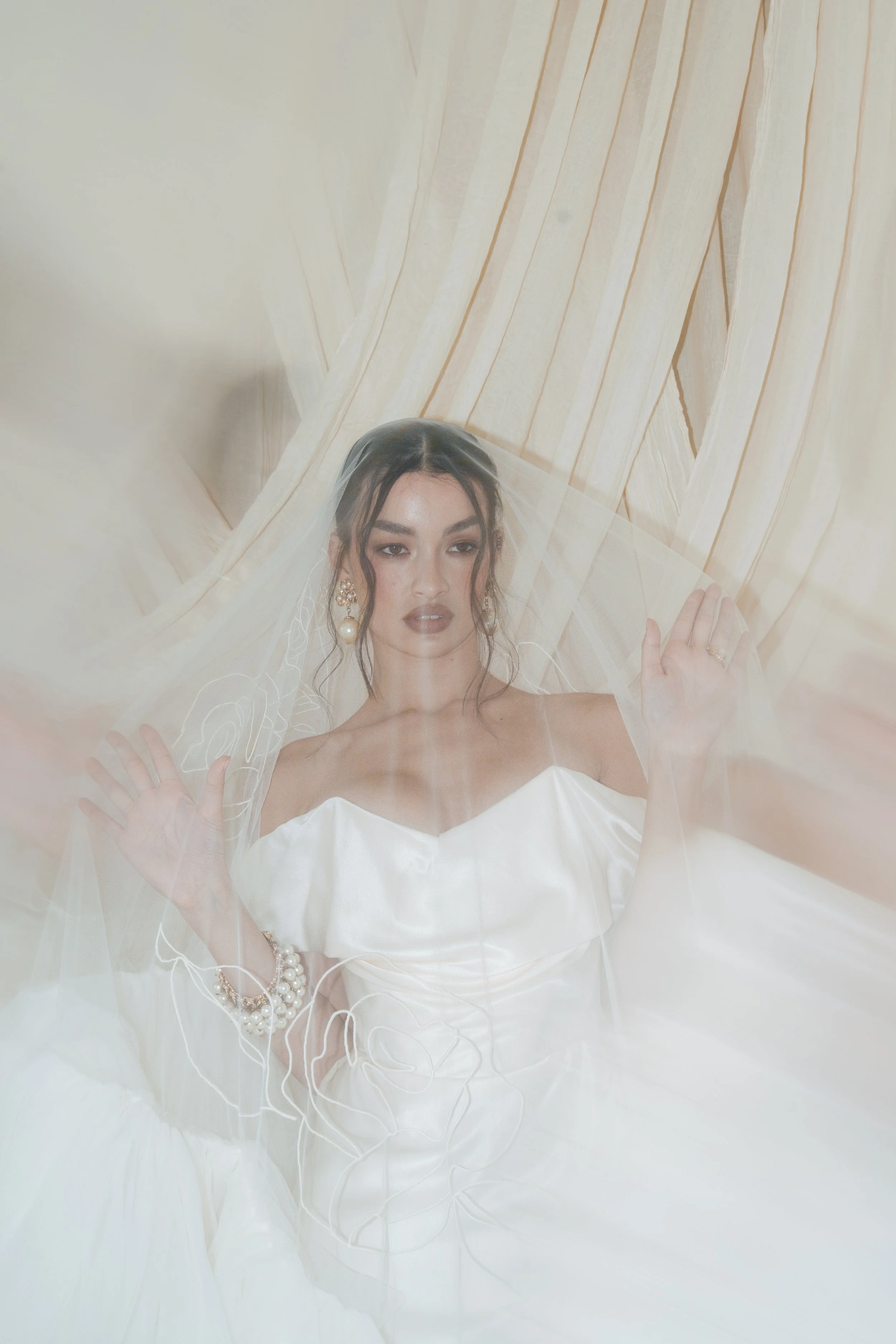 An ethereal, editorial bridal portrait of a woman in a Vivienne Westwood wedding dress and pearl jewellery, beautifully photographed from behind a sheer, draped lace veil.