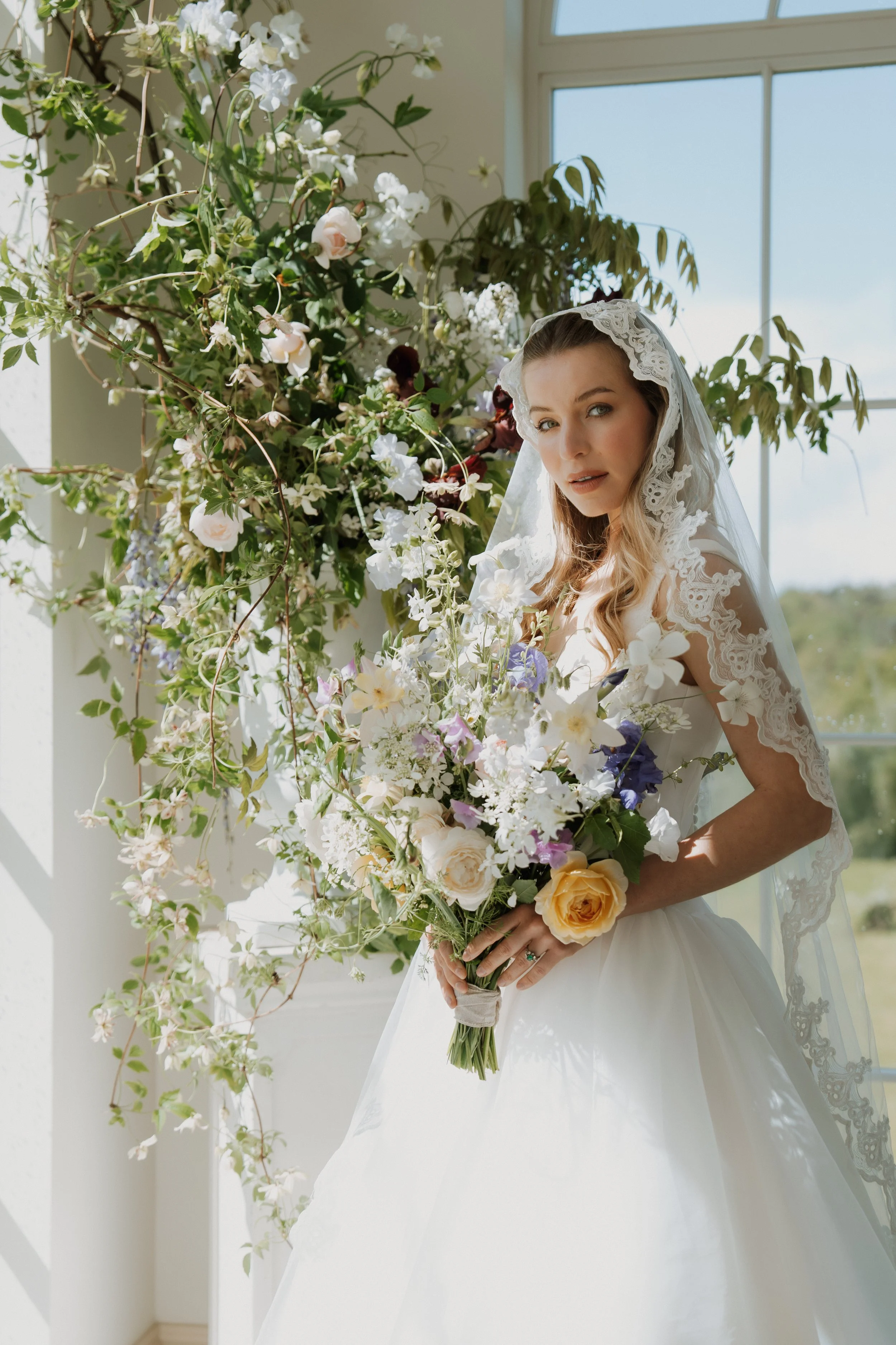 An elegant bride wearing a Savannah Miller Bridal wedding dress with lace sleeve details and a delicate lace veil, holding a vibrant bouquet near a large window.