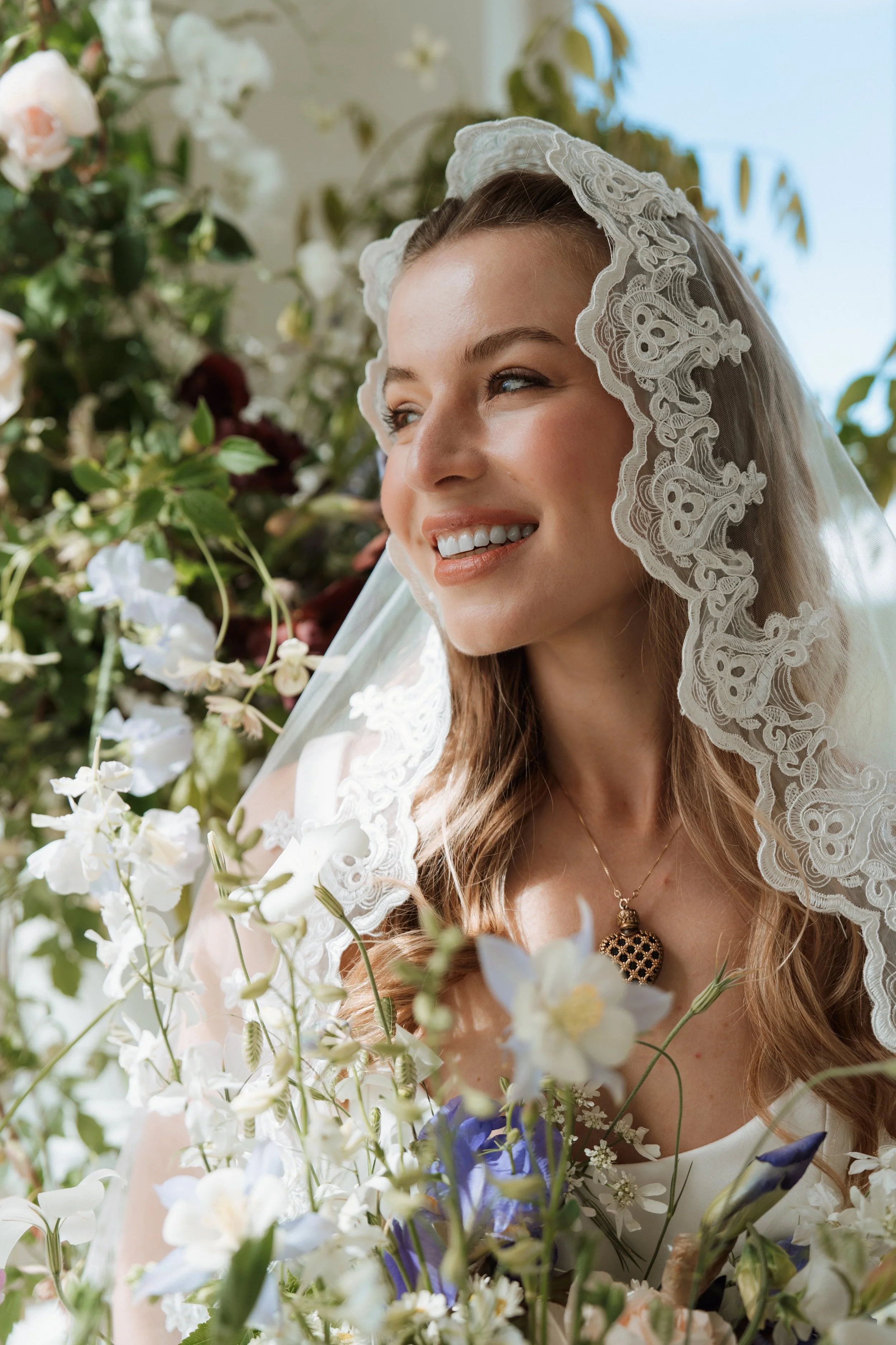 A smiling bride in a beautiful lace veil and wedding dress, surrounded by an abundance of elegant white and blue wedding flowers.