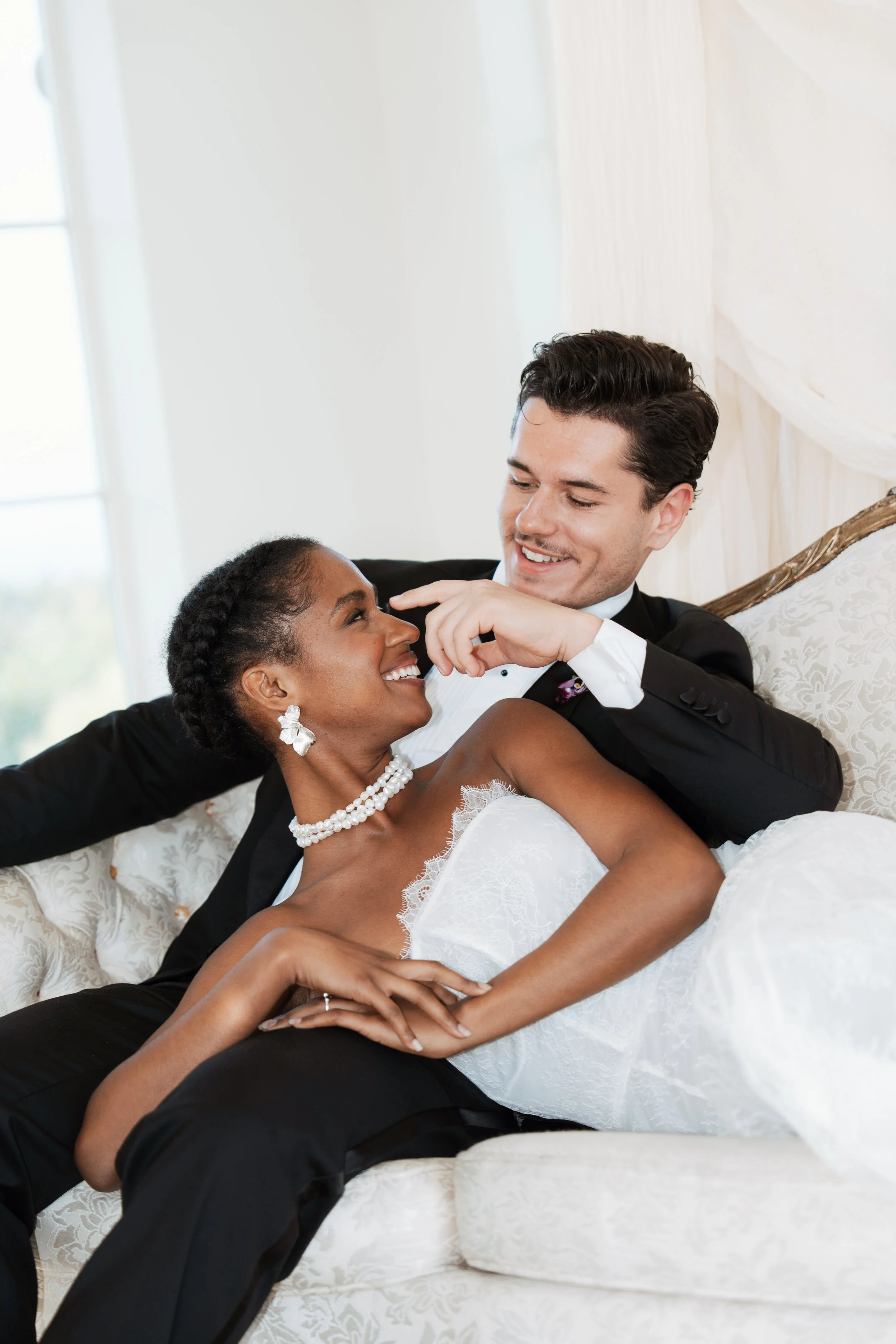 A newly married bride in a Vera Wang wedding gown and a groom in a classic tuxedo sitting on an ornate cream-colored couch, sharing an affectionate smile as he gently touches her face.