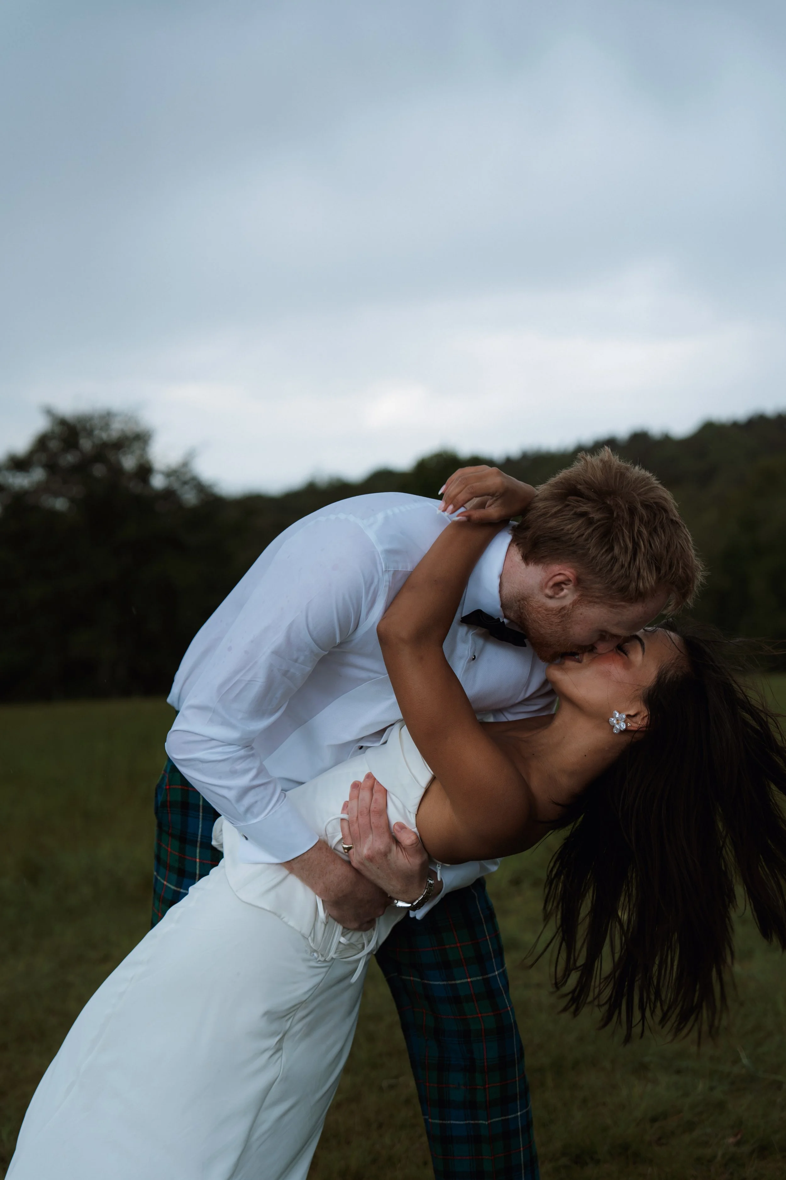 A joyful couple dancing outdoors on a cloudy day, featuring a groom in a traditional tartan and a smiling bride with black hair wearing an elegant Danielle Frankel wedding dress.