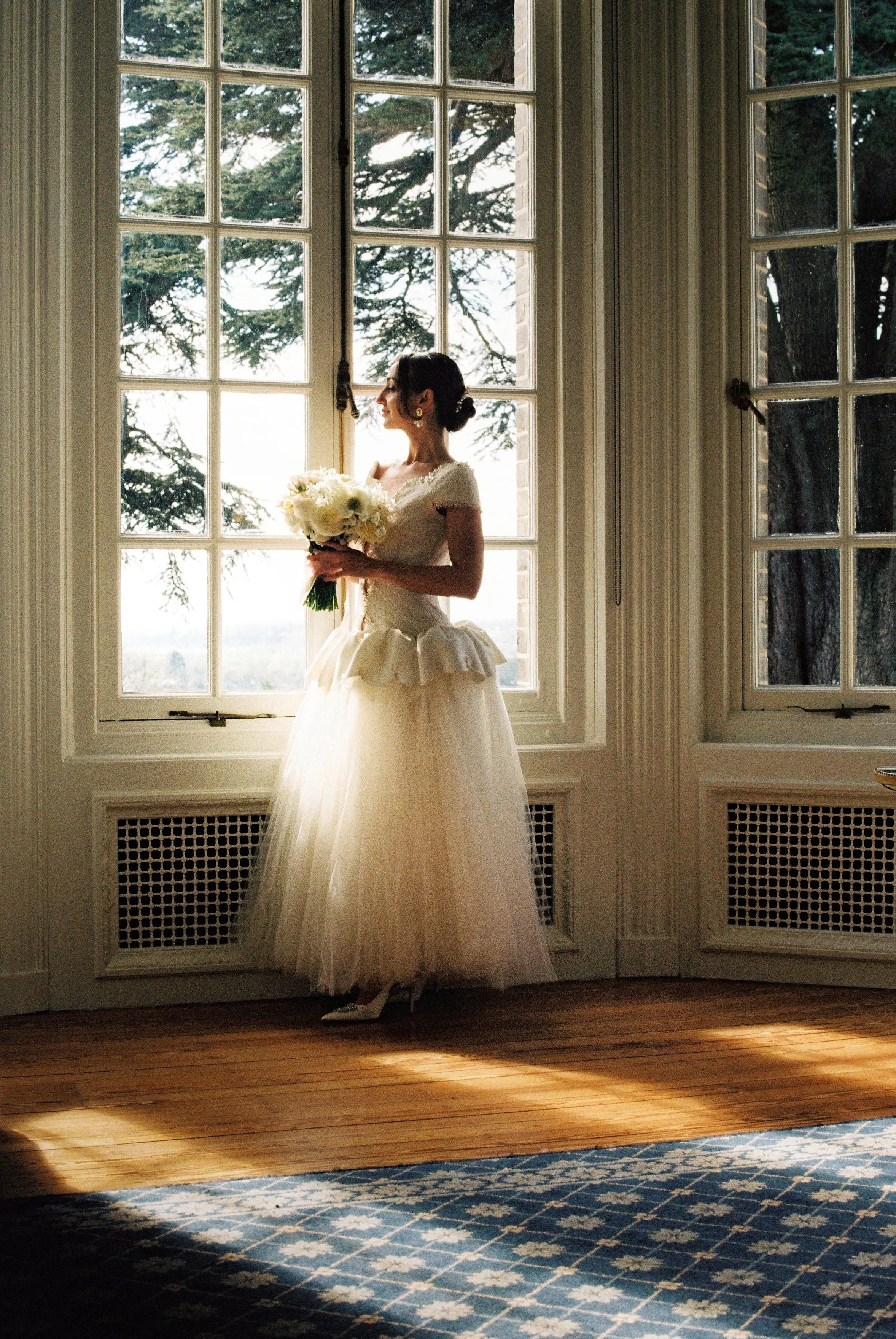 A bride in a white wedding dress holding a bouquet of flowers, standing by a large window with trees outside, in a room with wooden floors and radiator covers.