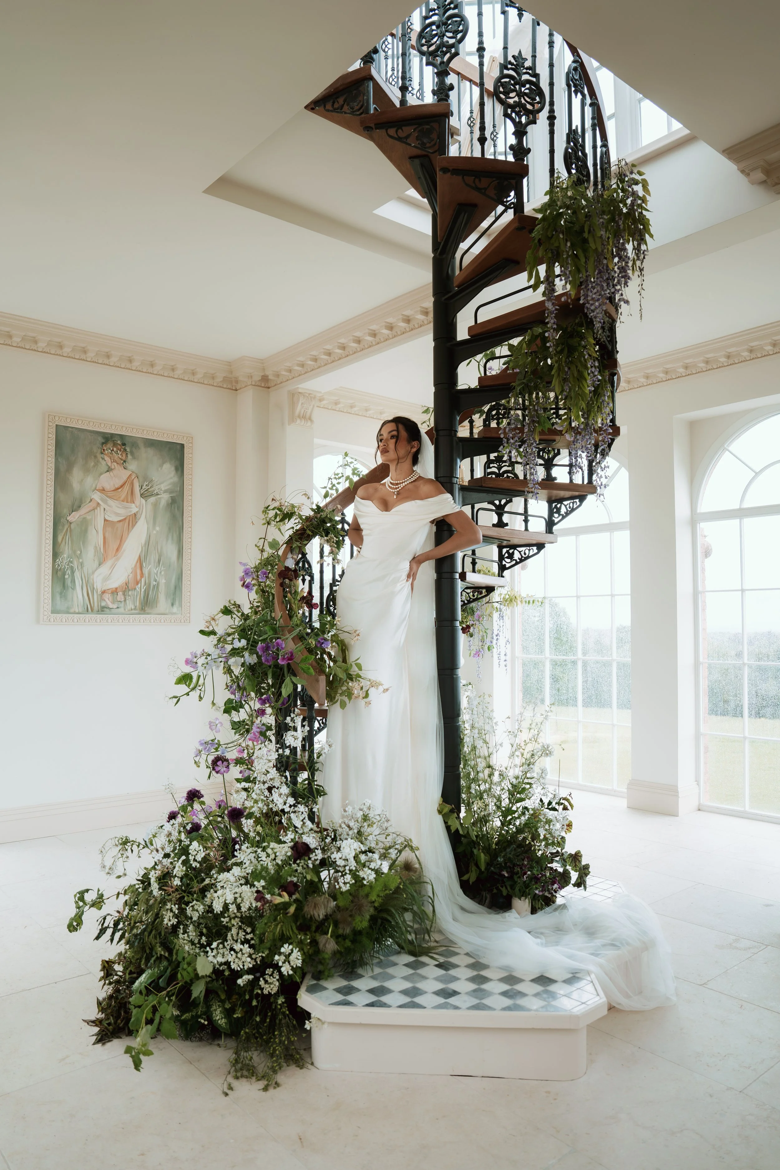 A woman in a white Vivienne Westwood wedding dress standing on a decorated staircase with black wrought iron railing in a bright room with large windows and floral arrangements.