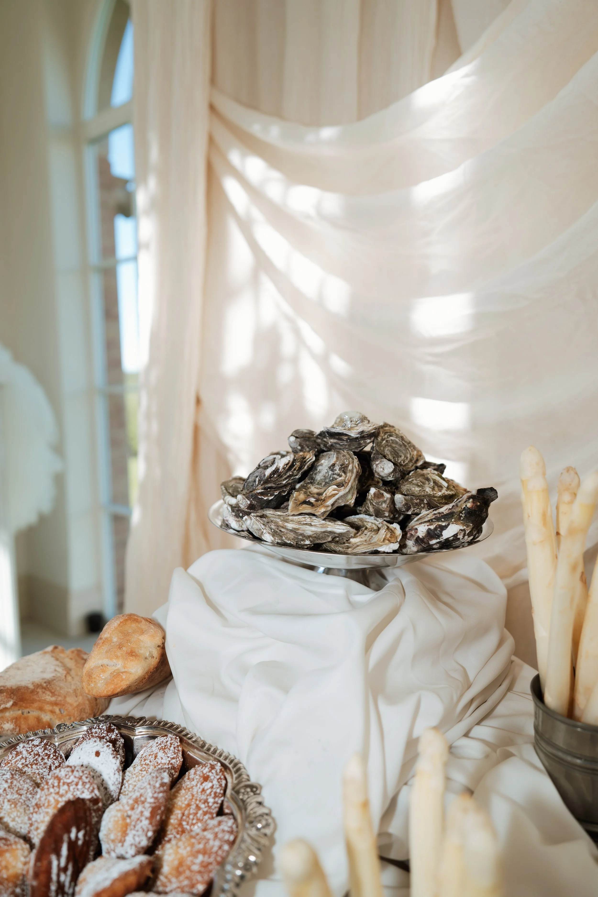 An elegant luxury wedding food station featuring fresh oysters, mussels, crusty bread, and white asparagus beautifully arranged on a table with white tablecloths.