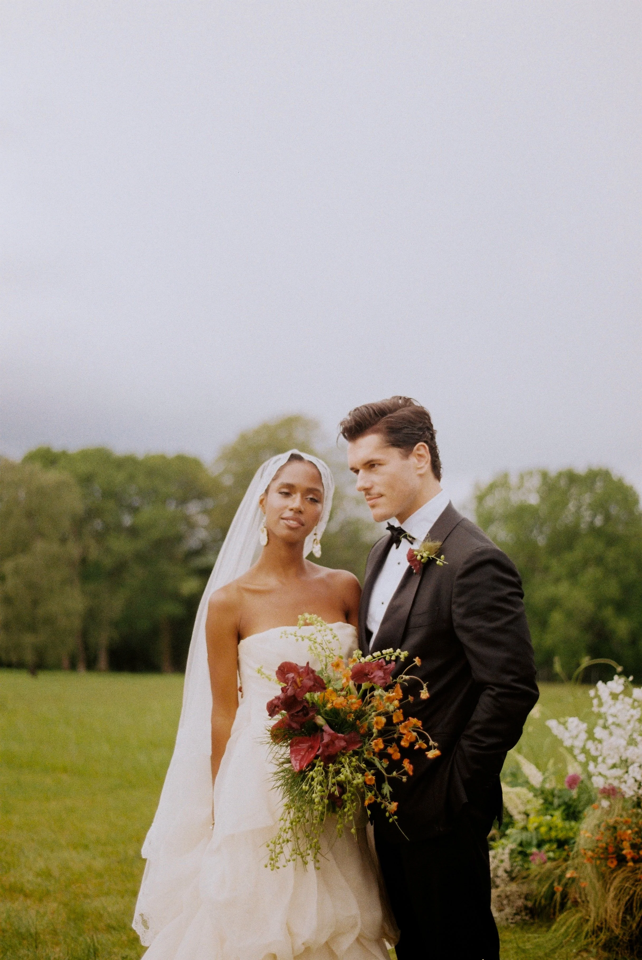 A bride and groom standing outdoors in a garden, dressed in wedding attire, with the bride holding a large bouquet of flowers and the groom in a black tuxedo, surrounded by greenery and colorful flowers.