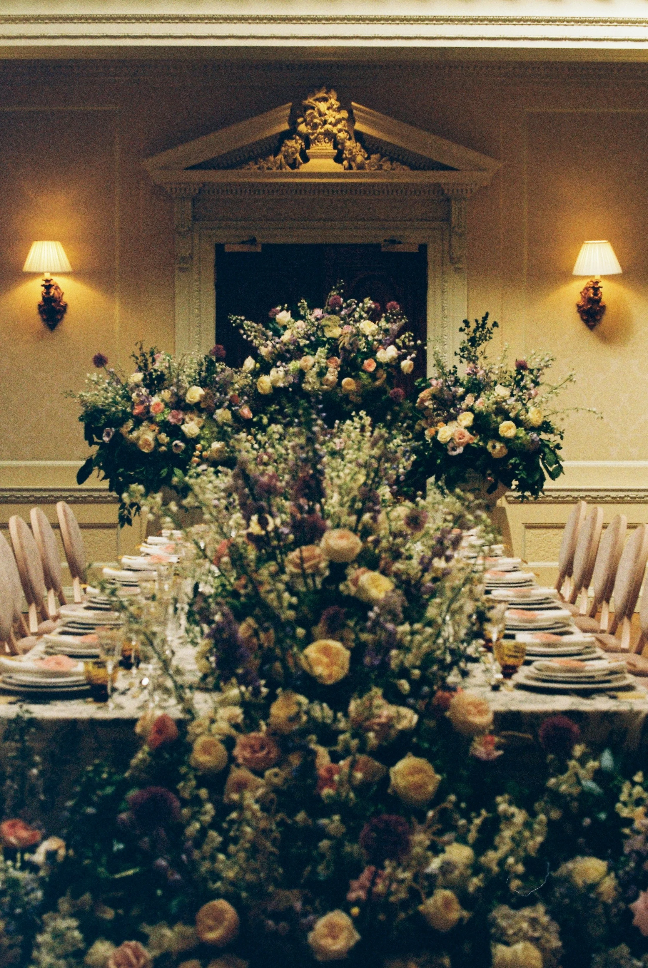 Decorated banquet table with a large floral centerpiece, set in an elegant room with wall-mounted lamps and ornate ceiling details at Hedsor House.
