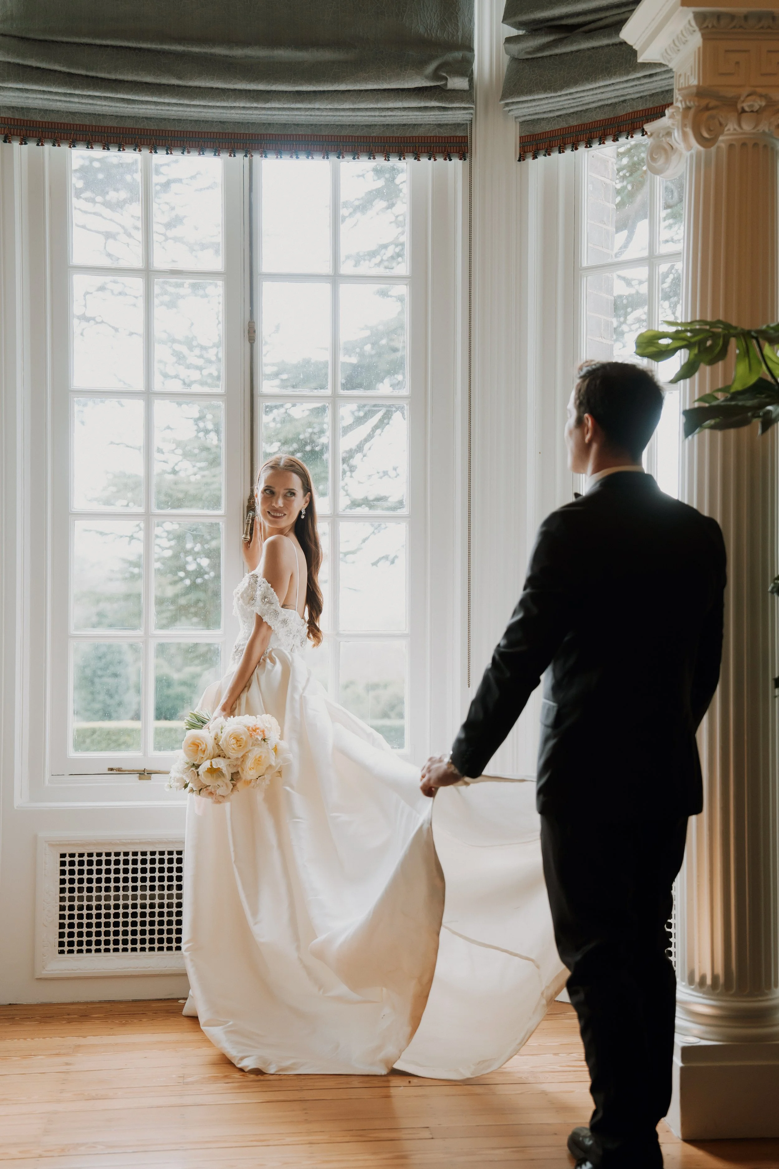 An elegant bride in a lace wedding gown holding hands with her groom in a tuxedo, standing by a grand window inside a luxury stateroom at Hedsor House.