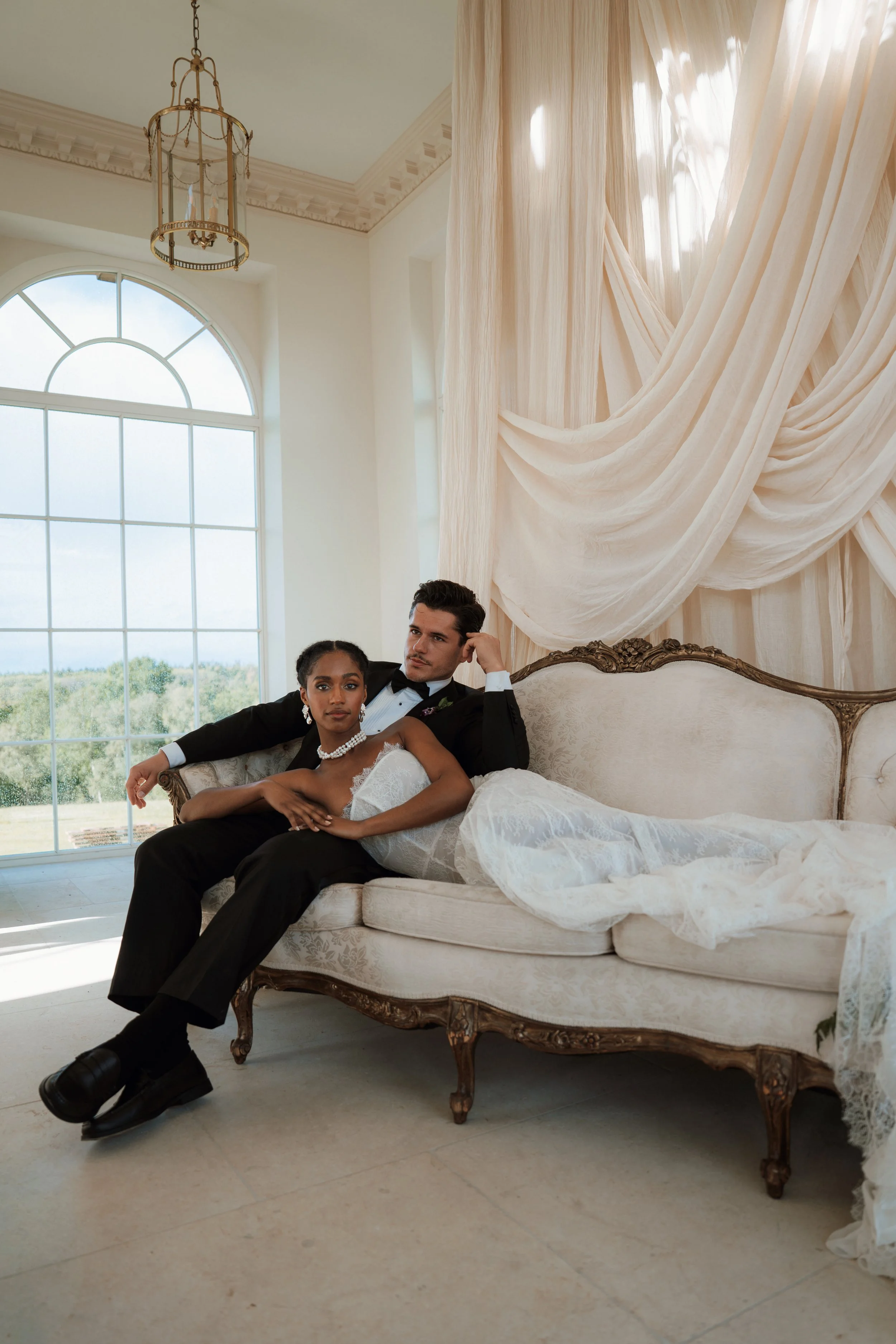 A happy bride and groom in elegant wedding attire lying on an ivory vintage couch in a bright room. The bride wears a luxury Asar London wedding dress with a pearl necklace and earrings, while the groom in a classic black tuxedo touches her face.