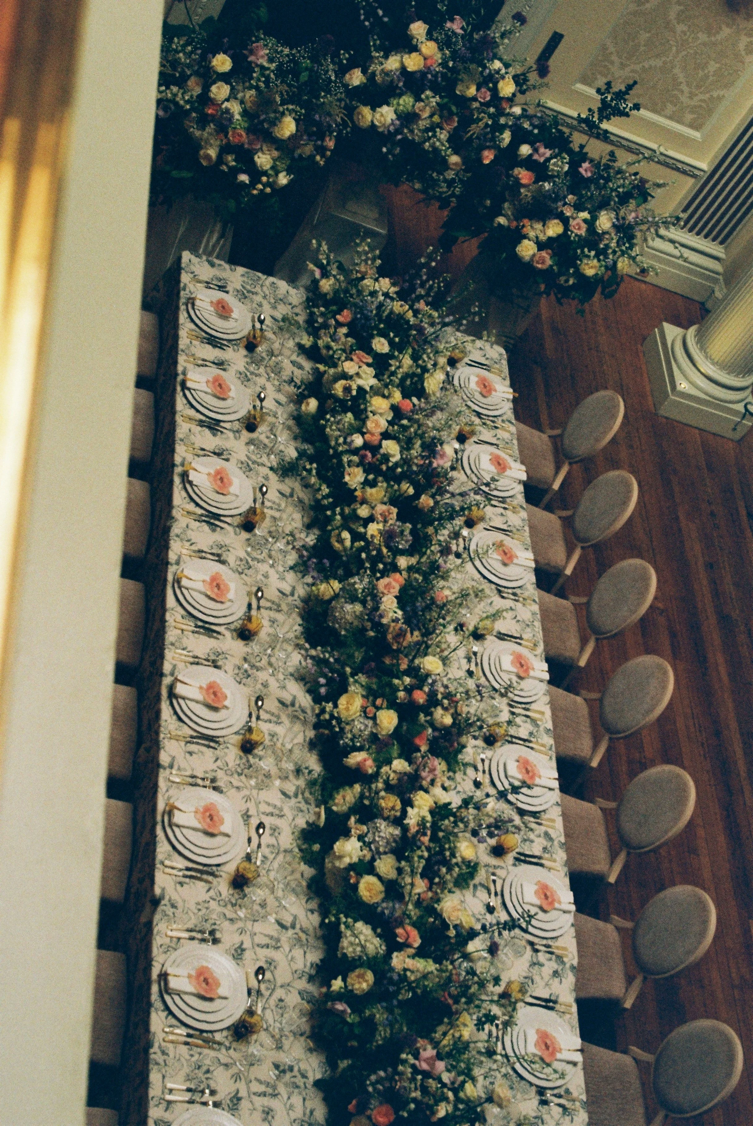 An elegant event table decorated with numerous floral arrangements and set for a formal dinner. The tablecloth has a floral pattern, and each place setting includes plates, utensils, and a small decorated napkin. Behind the table, there are more flow