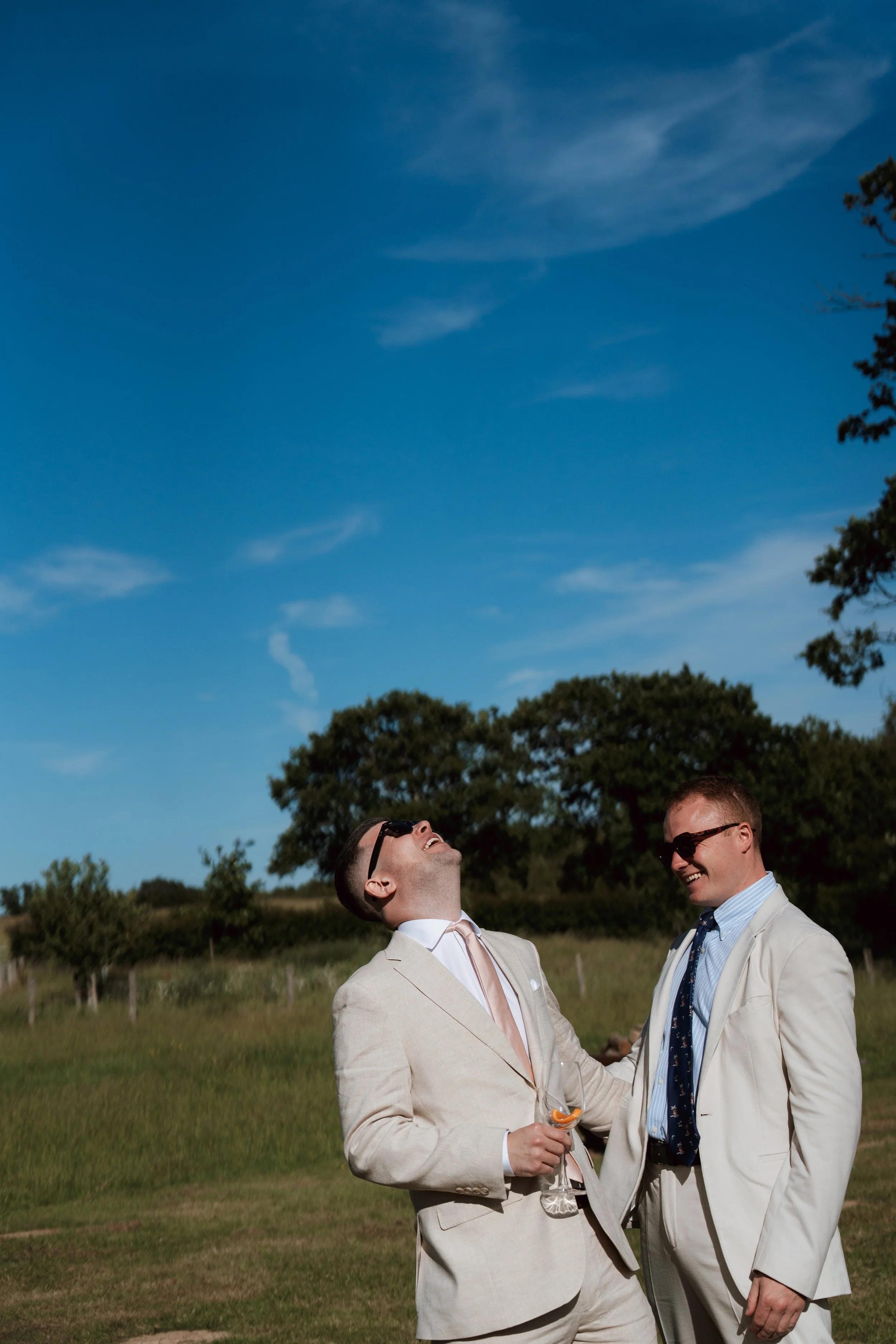 Two wedding guests laughing together outdoors beneath a vivid blue sky