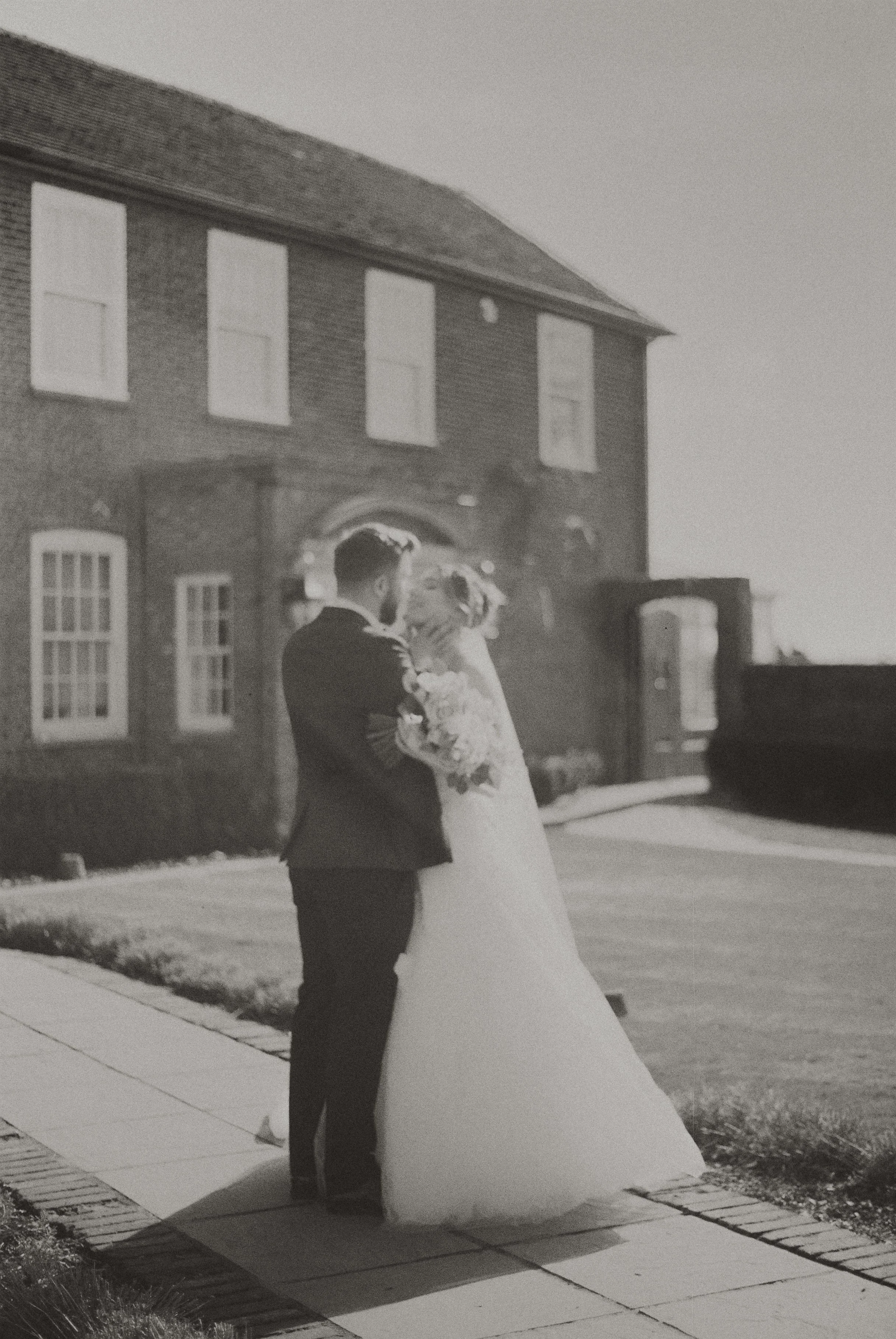 A bride and groom standing on a sidewalk outside a brick building, embracing on their wedding day with a large wedding dress and tuxedo.