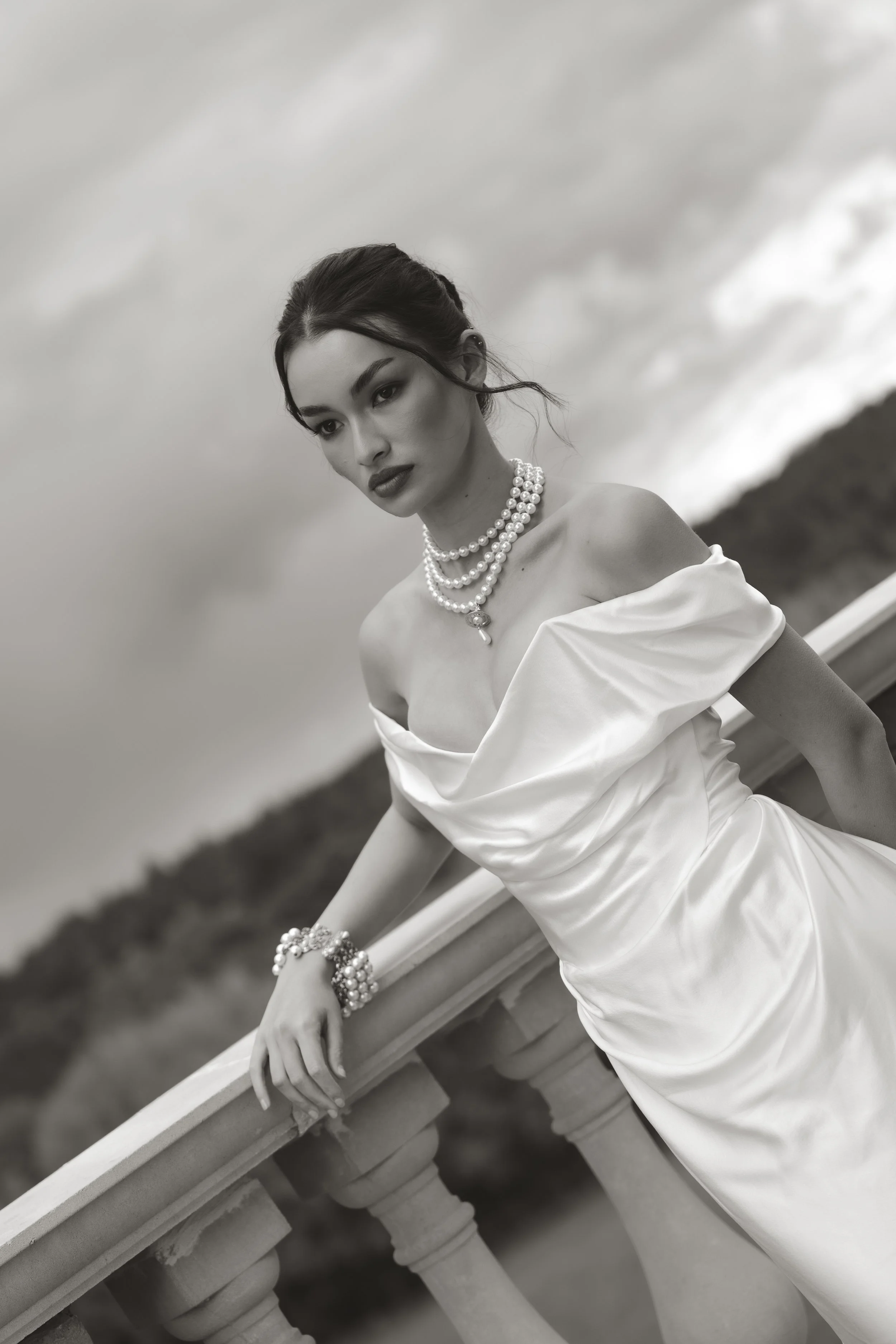Black and white editorial wedding portrait of an elegant bride wearing a strapless satin Vivienne Westwood wedding dress, styled with multiple pearl necklaces and bracelets, leaning on an outdoor balustrade against a cloudy sky.