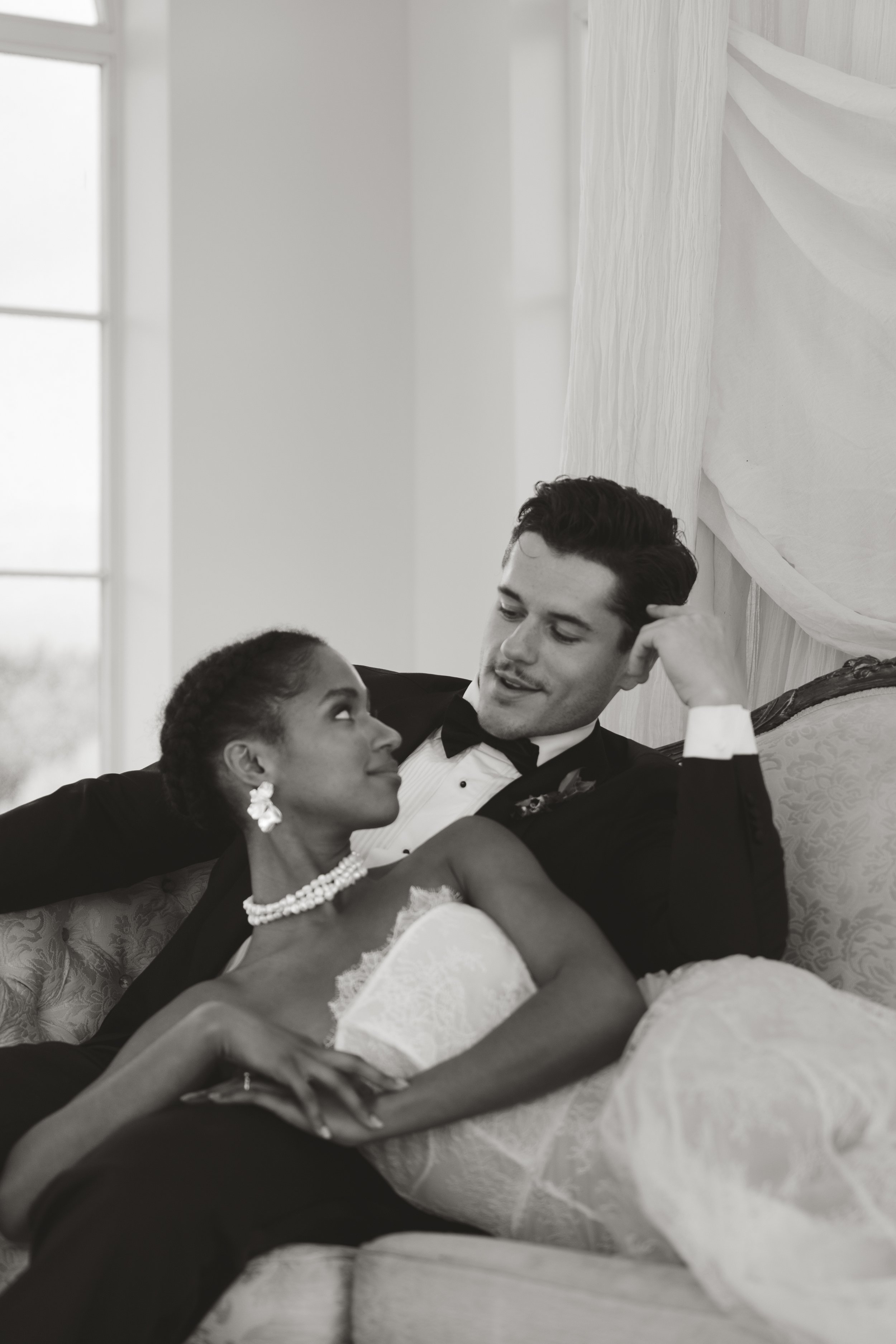 Black and white editorial wedding portrait of a couple lounging on a sofa in a bright room; the bride wears a strapless Vera Wang wedding dress styled with pearl jewelry while gazing at her groom in a tuxedo.