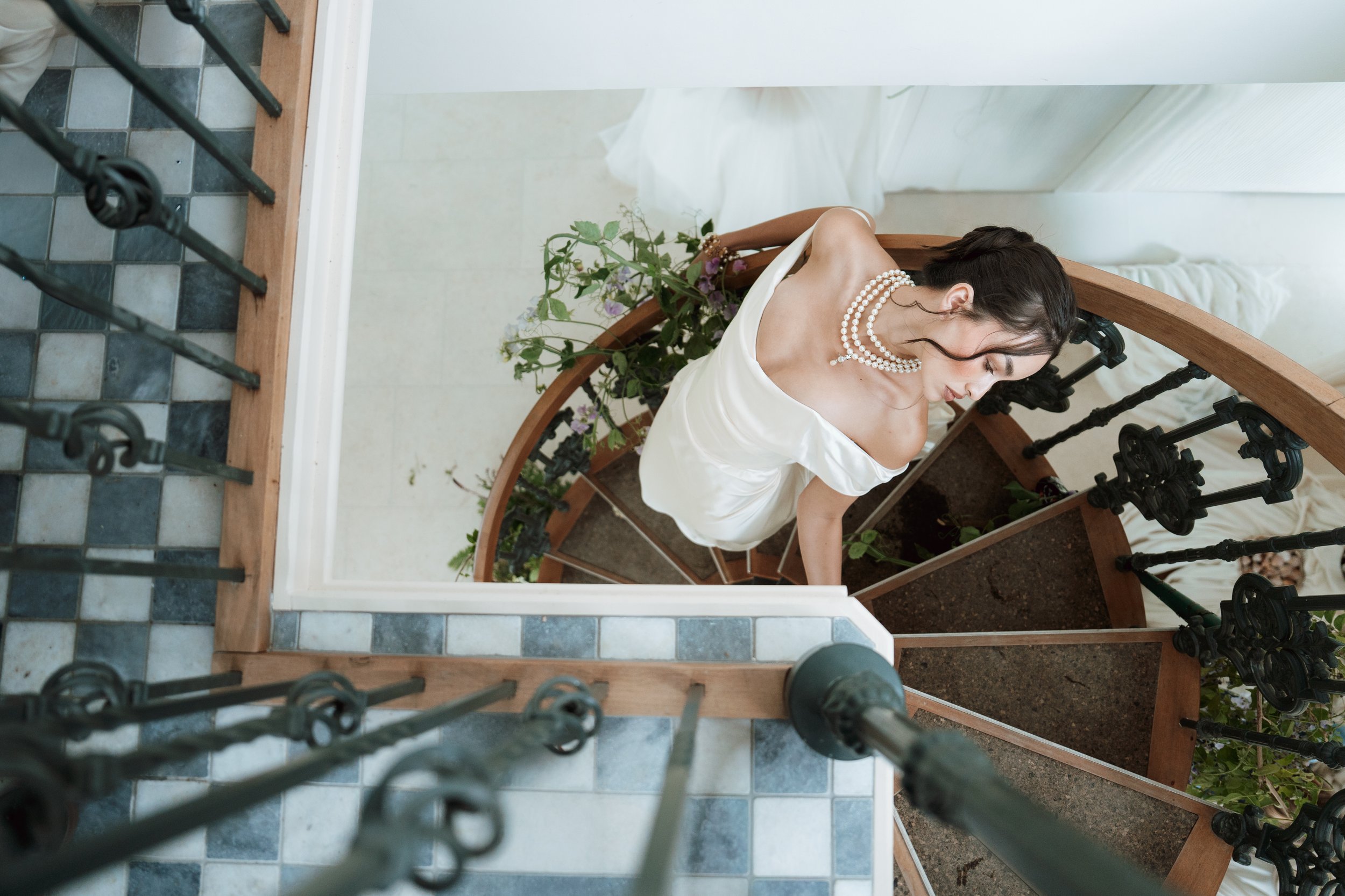 A bride in Vivienne Westwood dress and pearl necklace standing on a spiral staircase with wooden steps and black wrought iron railing, holding a bouquet of flowers.